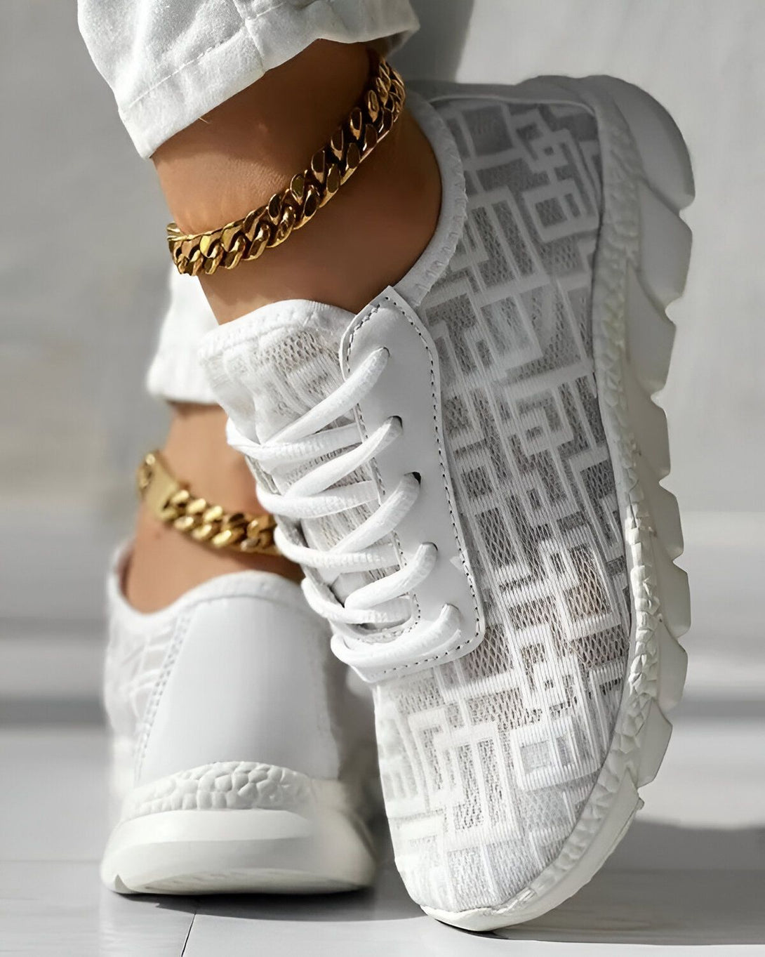 Une femme porte des baskets blanches avec des lacets blancs et une semelle blanche. Les baskets sont en maille et ont un motif de monogramme. La femme porte également un pantalon blanc et un bracelet de cheville en or.