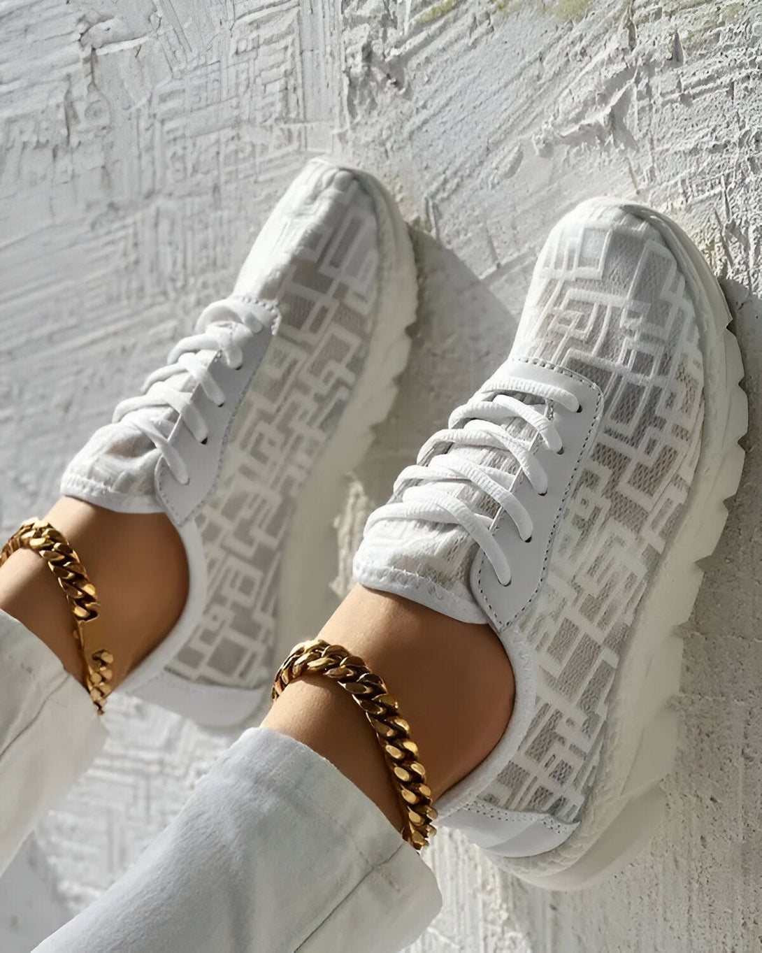 Une paire de baskets blanches avec des lacets blancs et une semelle blanche. Les baskets sont portées par une femme qui porte un pantalon blanc et des chaussettes blanches. La femme est assise sur un mur blanc.