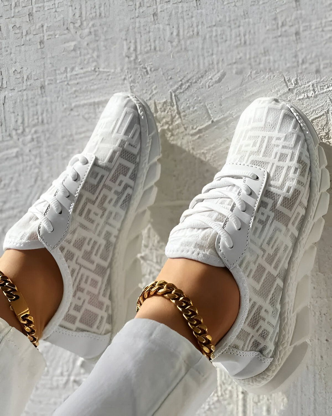 Une paire de baskets blanches avec un motif de monogramme Fendi. Les baskets sont portées par une femme qui porte également un pantalon blanc et un bracelet de cheville en or.
