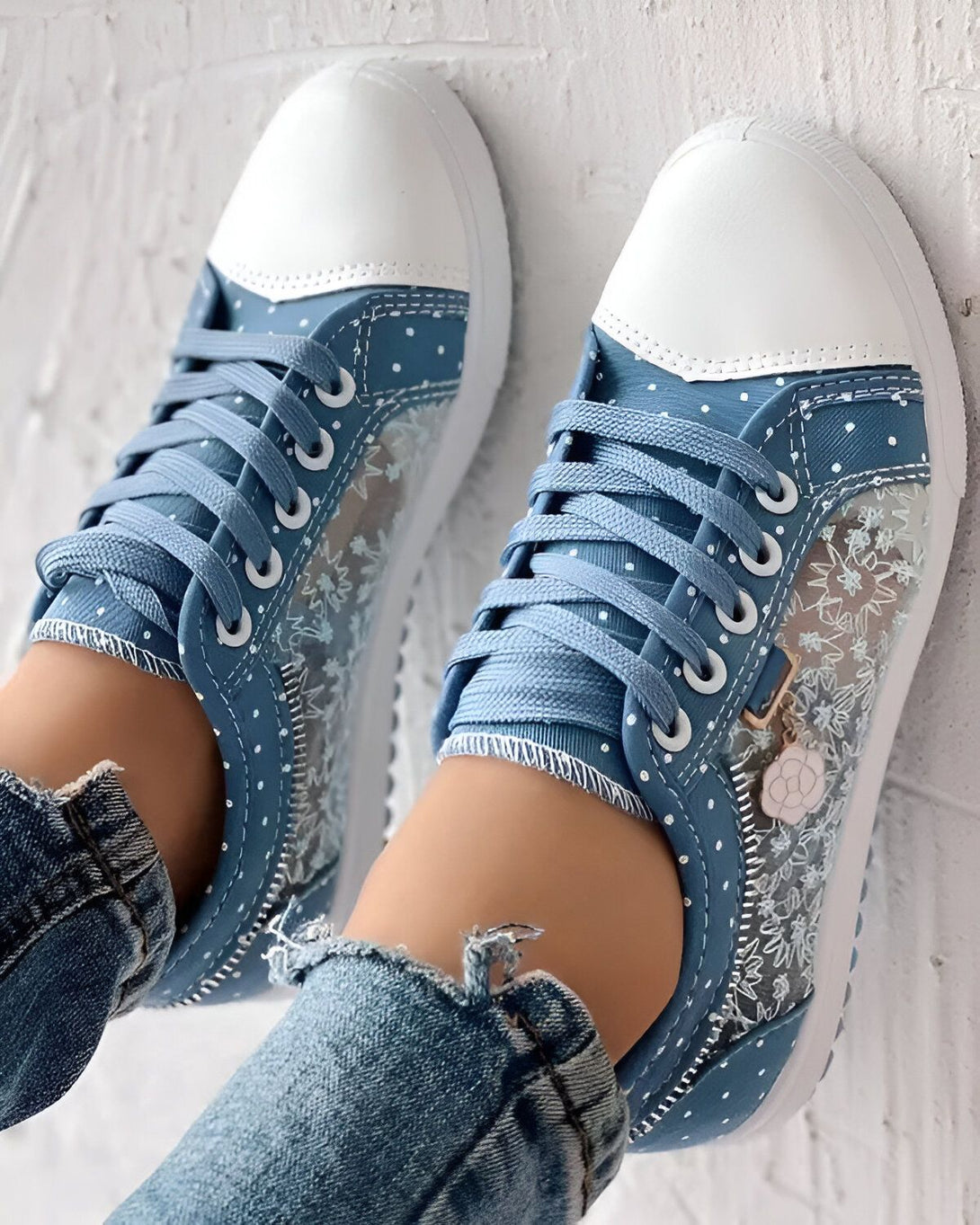 Une paire de baskets bleues et blanches avec un motif floral transparent sur le côté. Les baskets sont lacées et ont une fermeture éclair sur le côté. Elles sont portées avec un jean bleu troué.