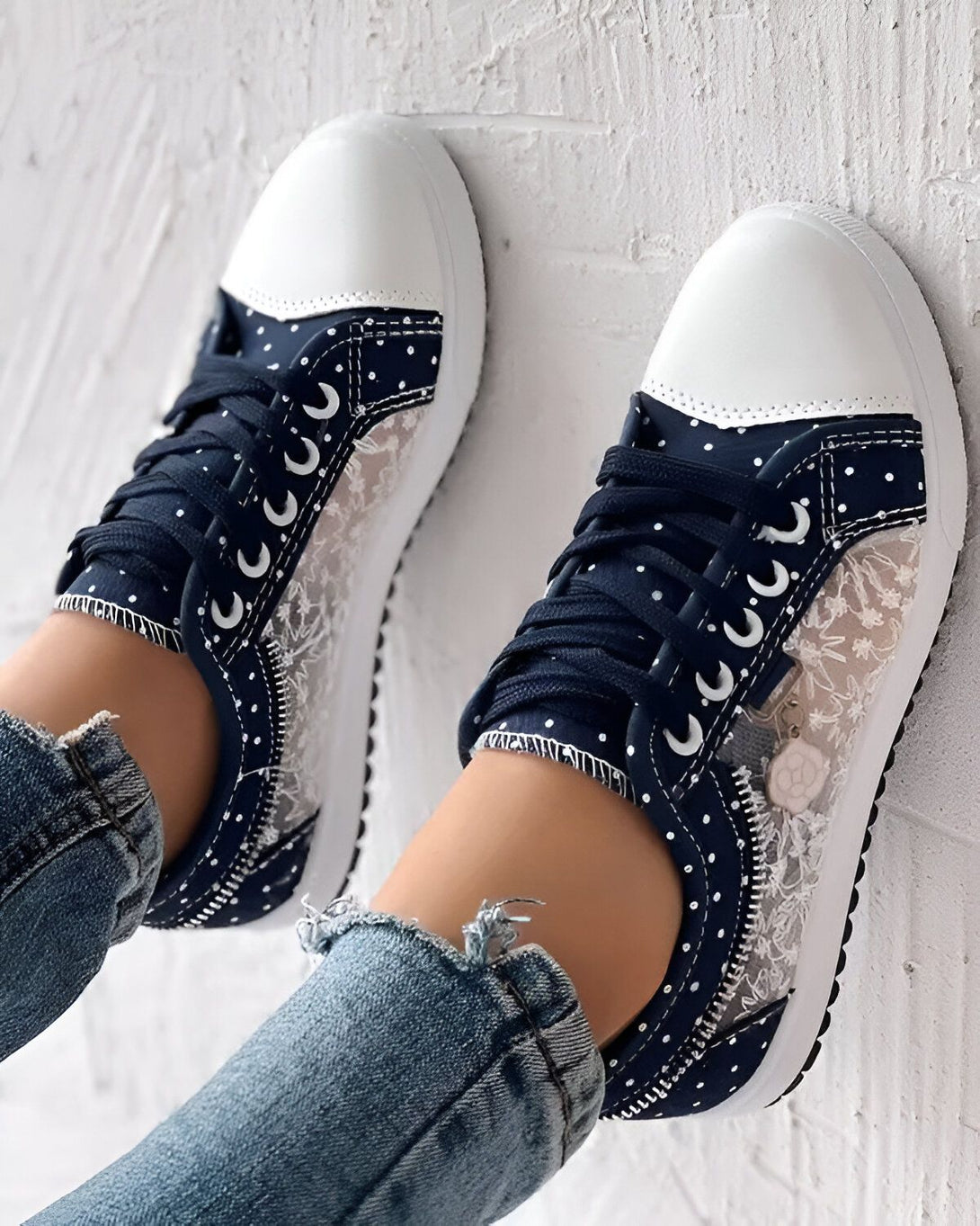 Une paire de baskets basses bleu marine à lacets avec un empiècement en dentelle blanche sur le côté. Les baskets sont portées avec un jean bleu troué.