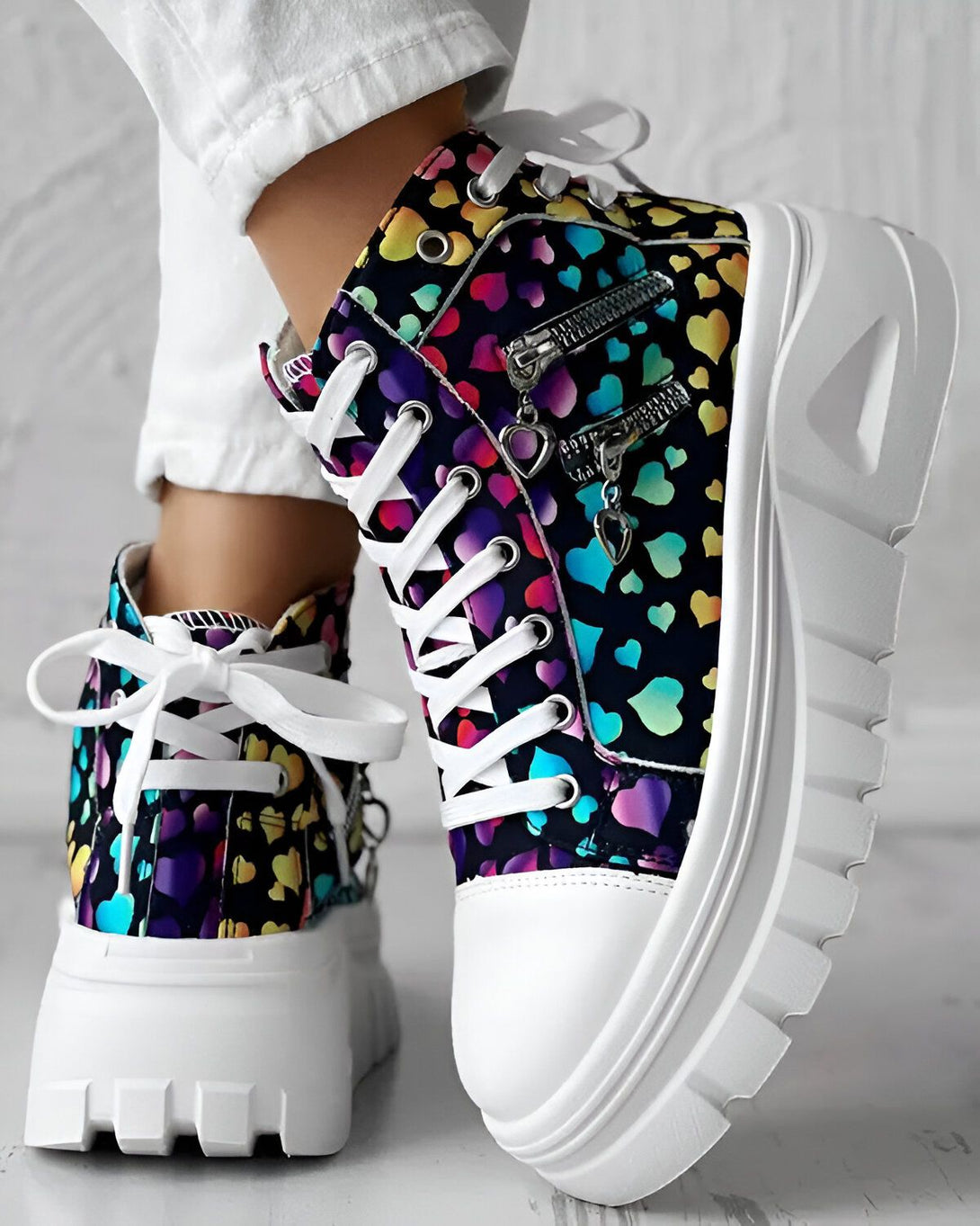Une paire de chaussures montantes à lacets avec un motif de cœurs multicolores sur fond noir. Les semelles sont blanches et épaisses.
