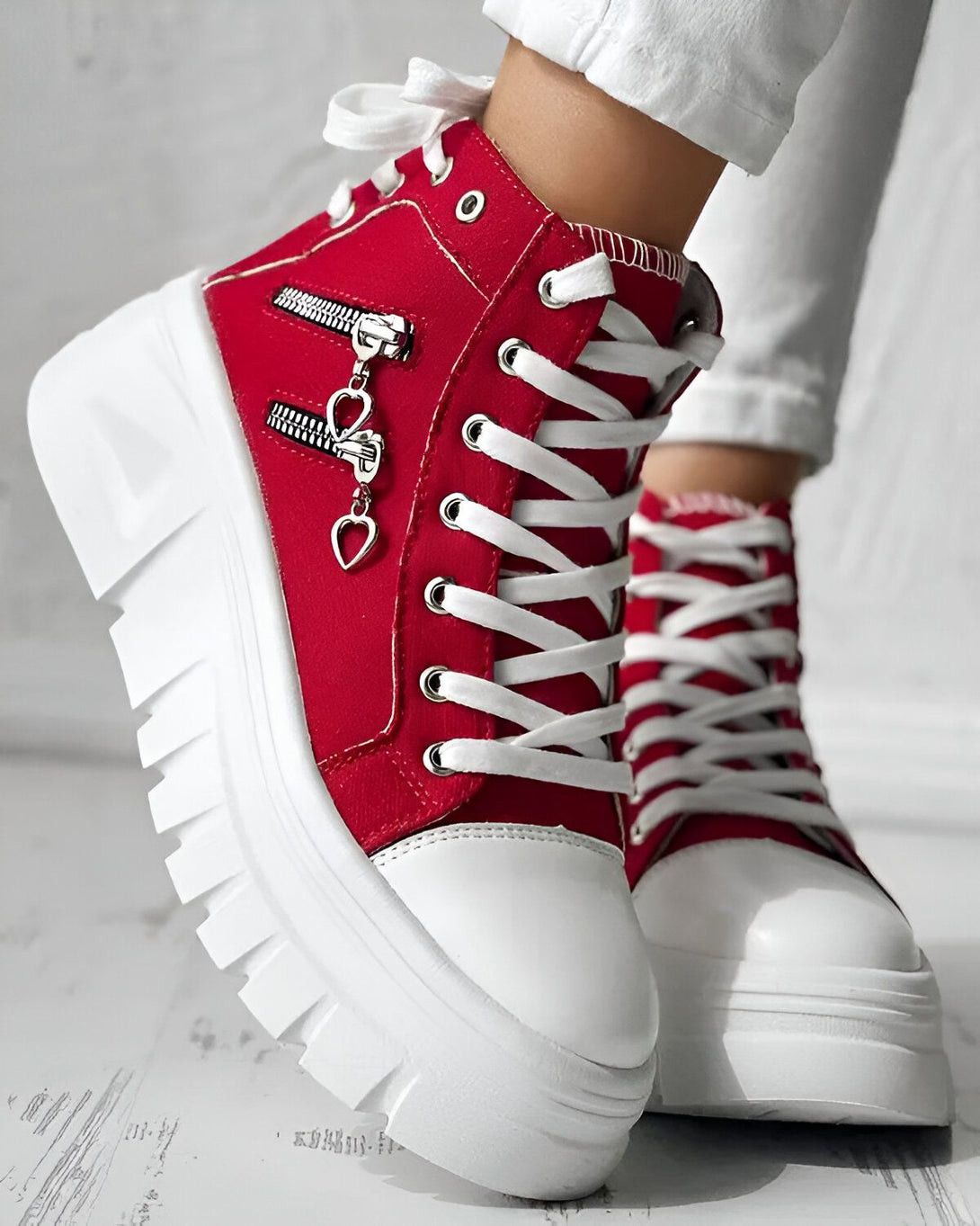 Une paire de bottes rouges à lacets avec une fermeture éclair sur le côté et un cœur en métal sur la fermeture éclair. Les semelles des bottes sont blanches.