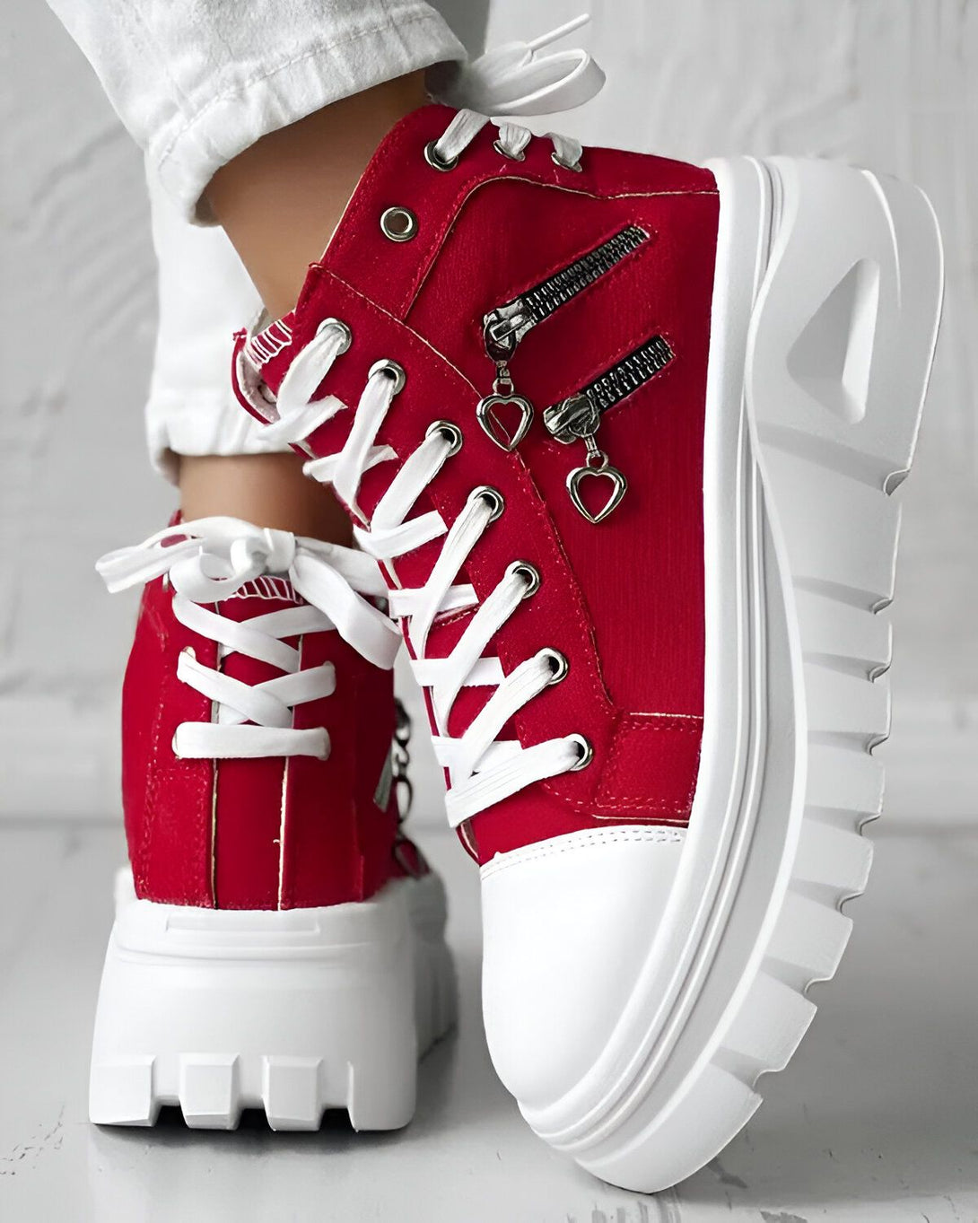 Une paire de bottes rouges à lacets avec une fermeture éclair sur le côté. Les bottes ont une semelle blanche épaisse et des cœurs en métal sur les fermetures éclair.