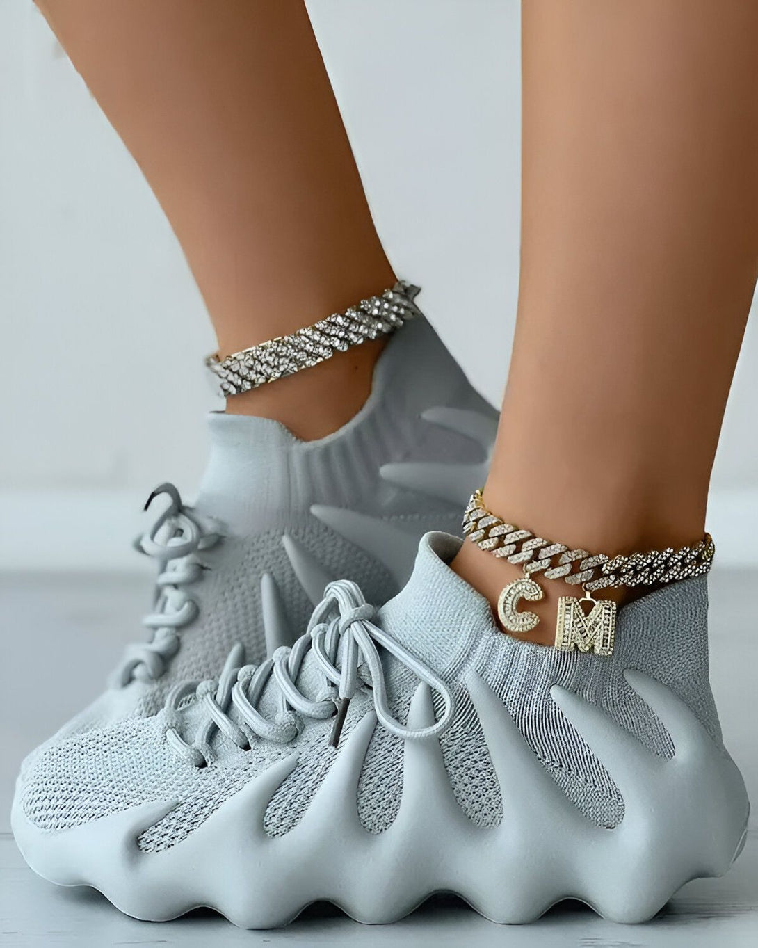 Une femme porte des baskets grises avec des lacets gris et une semelle épaisse. Elle porte également un bracelet de cheville en or avec des lettres en diamant.
