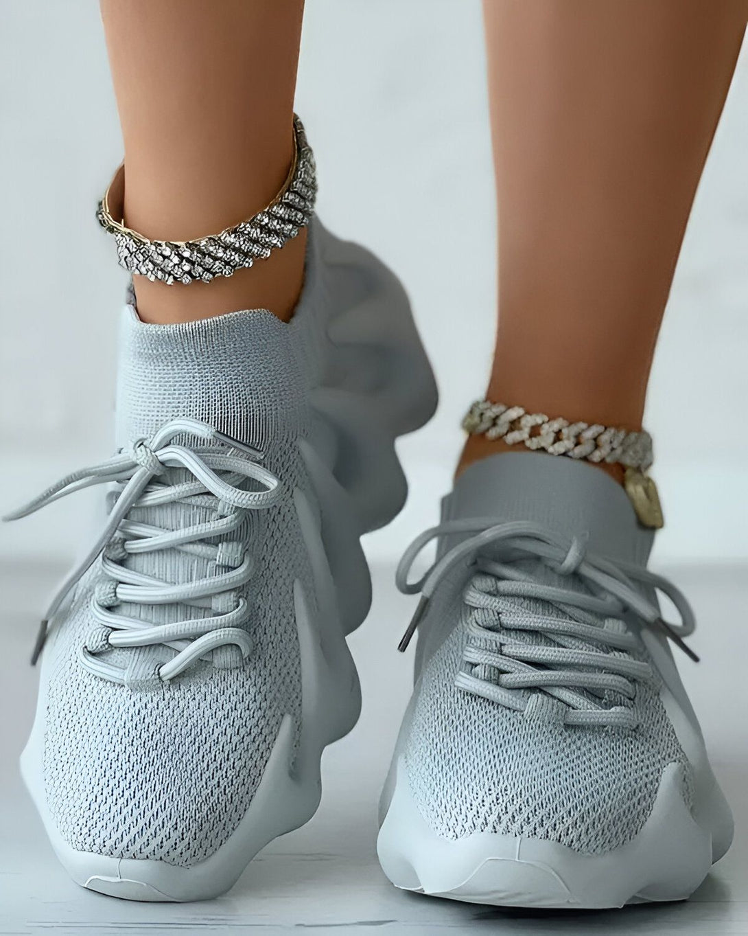 Une paire de baskets grises à lacets avec une semelle épaisse et une tige en maille. Les baskets sont portées par une femme qui a une chaîne en or à la cheville.