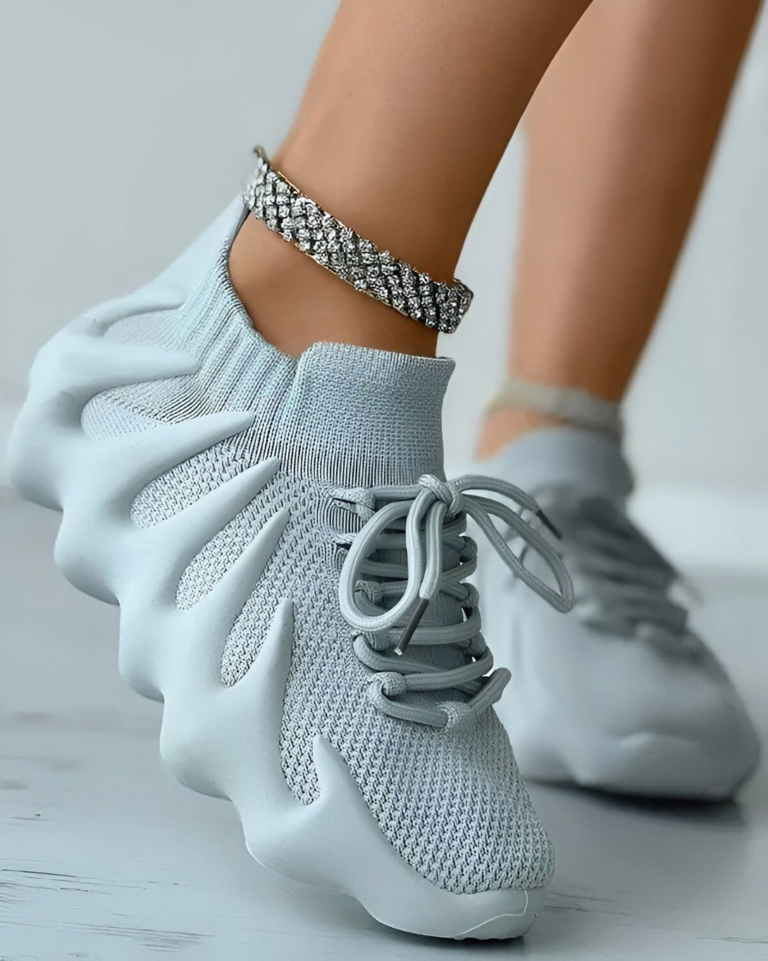 Une femme porte des baskets grises avec des lacets gris et une semelle épaisse. Elle porte également un bracelet de cheville en diamant.