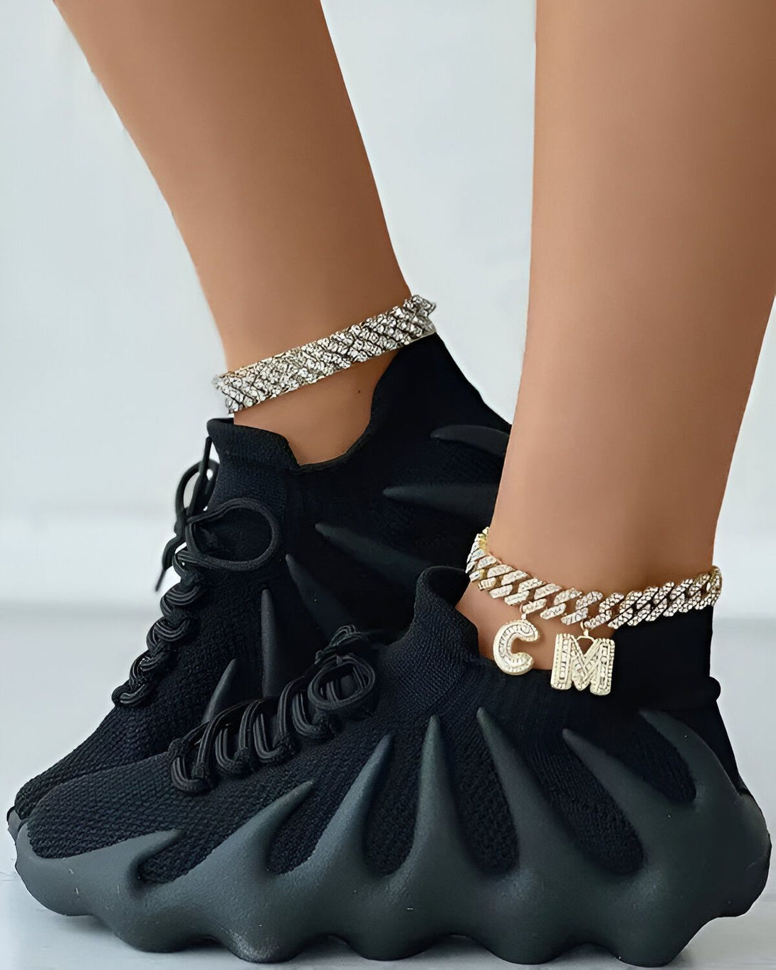 Une femme porte des baskets noires avec des lacets noirs et des semelles épaisses. Elle porte également deux bracelets de cheville en argent et or avec des pendentifs.