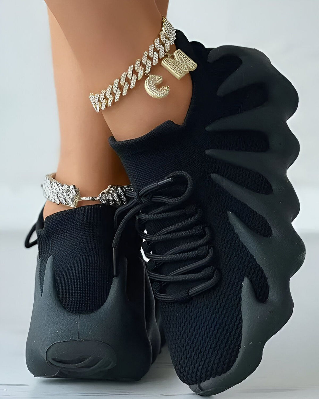 Une femme porte des baskets noires avec des lacets noirs et une grosse semelle. Elle porte également un bracelet de cheville en or avec un pendentif en forme de lettre C.