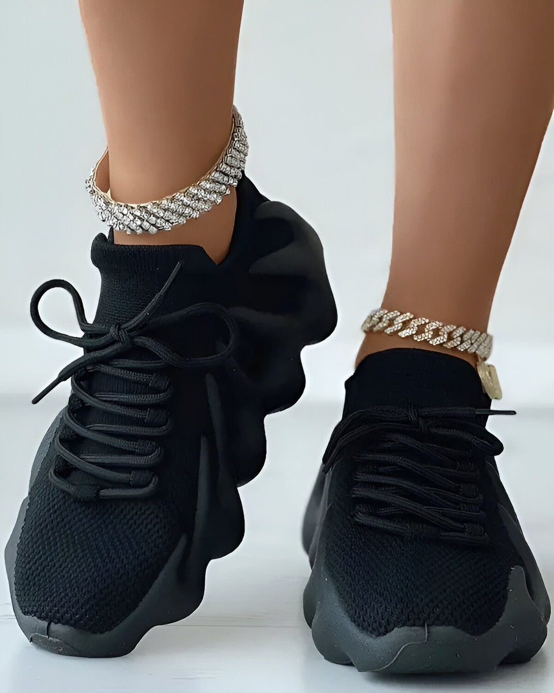 Une paire de baskets noires avec des lacets noirs et une semelle épaisse. Les baskets sont portées par une femme qui a des bracelets de cheville en diamant.