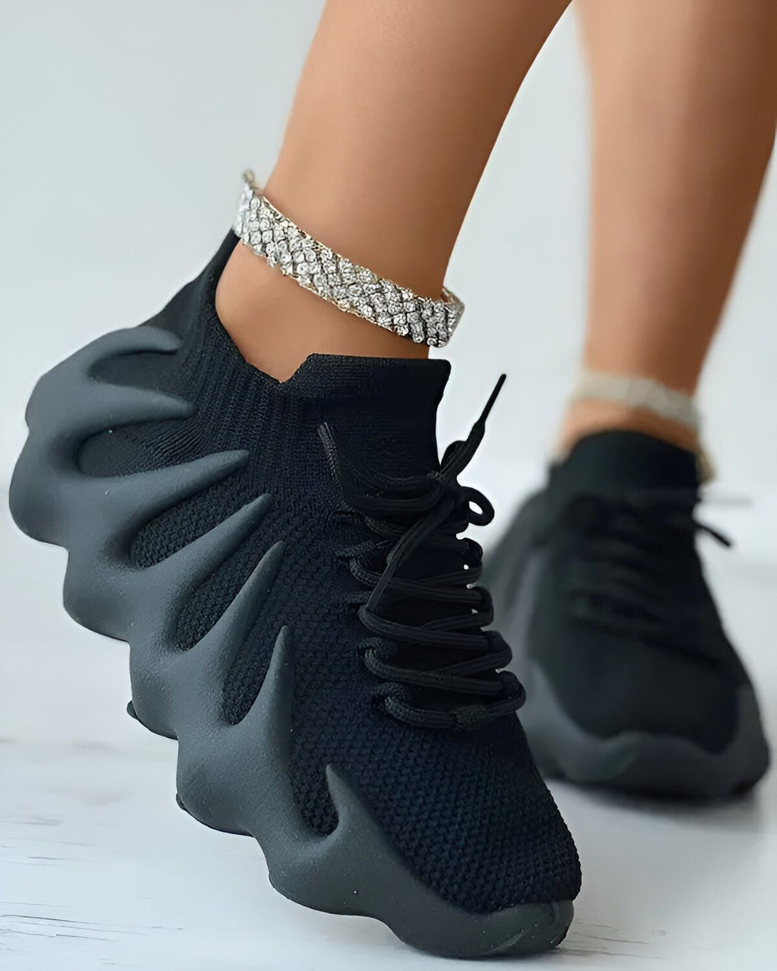 Une femme porte des baskets noires avec des lacets noirs et une semelle épaisse. Elle porte également un bracelet de cheville en diamant.