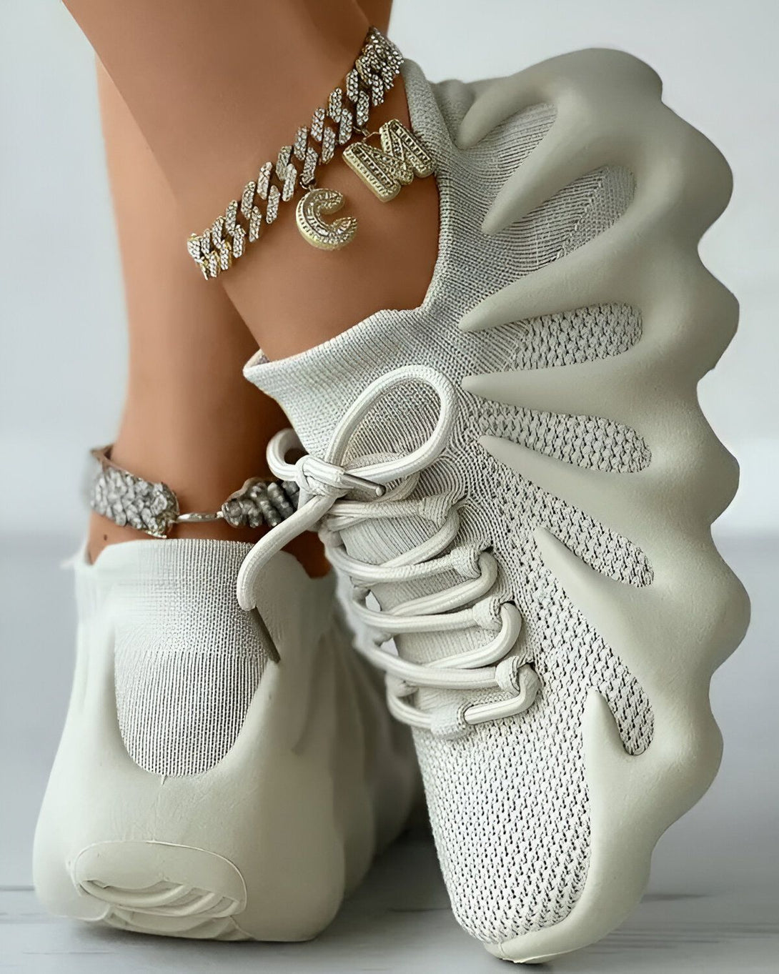Une femme porte des baskets blanches avec des lacets blancs. Elle porte également un bracelet de cheville en or avec un pendentif en forme de lettre "C".