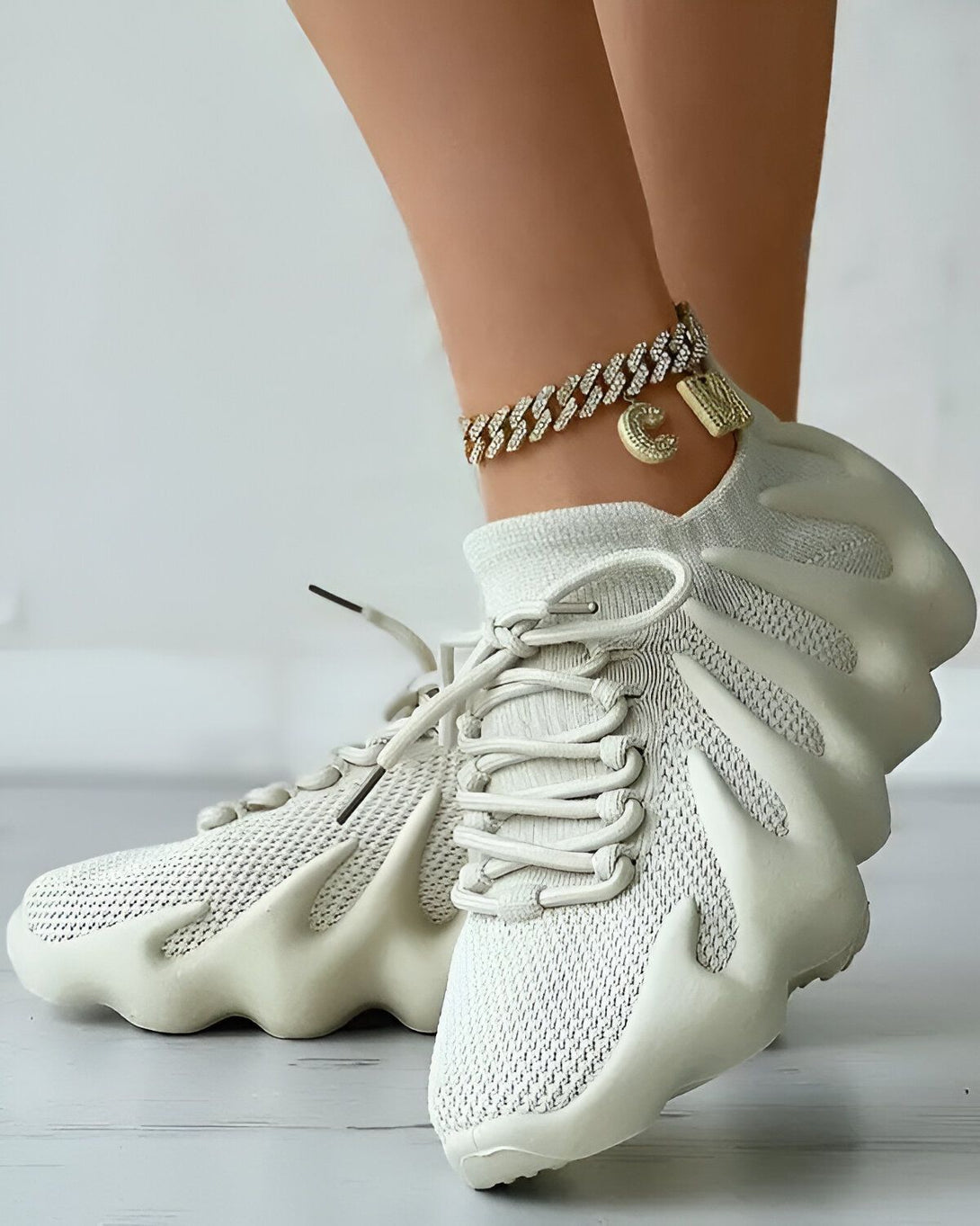 Une femme porte des baskets blanches avec un lacet blanc et une semelle épaisse. Elle porte également un bracelet de cheville en or avec un pendentif en forme de lettre C.