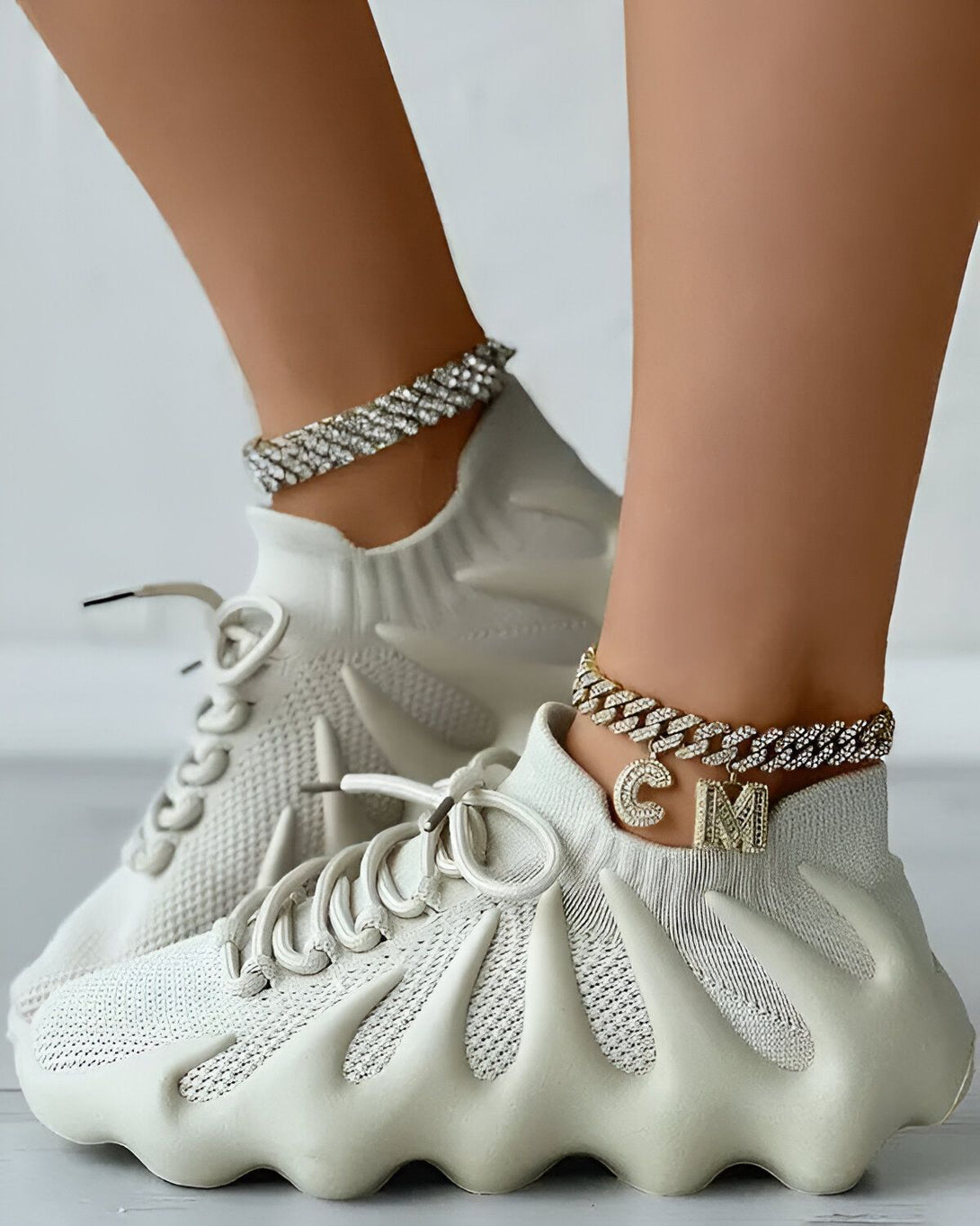 Une femme porte des baskets blanches avec des lacets blancs et une chaîne de cheville en or avec un pendentif en forme de lettre "C" et "M".