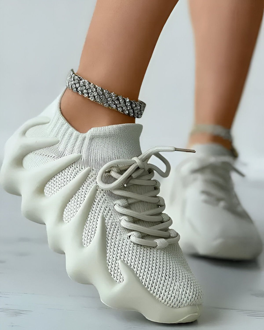 Une femme porte des baskets blanches avec des lacets blancs et une semelle épaisse. Elle porte également un bracelet de cheville en strass.
