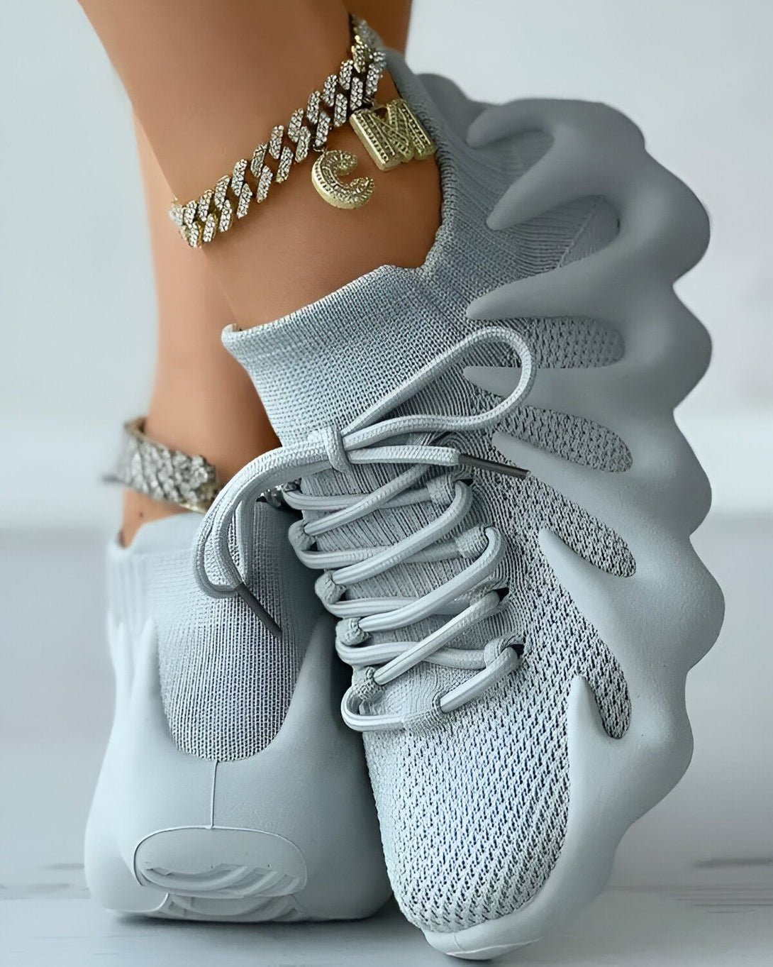 Une femme porte des baskets grises avec des lacets gris et une semelle blanche. Elle porte également un bracelet de cheville en or avec un pendentif en forme de lettre "M".