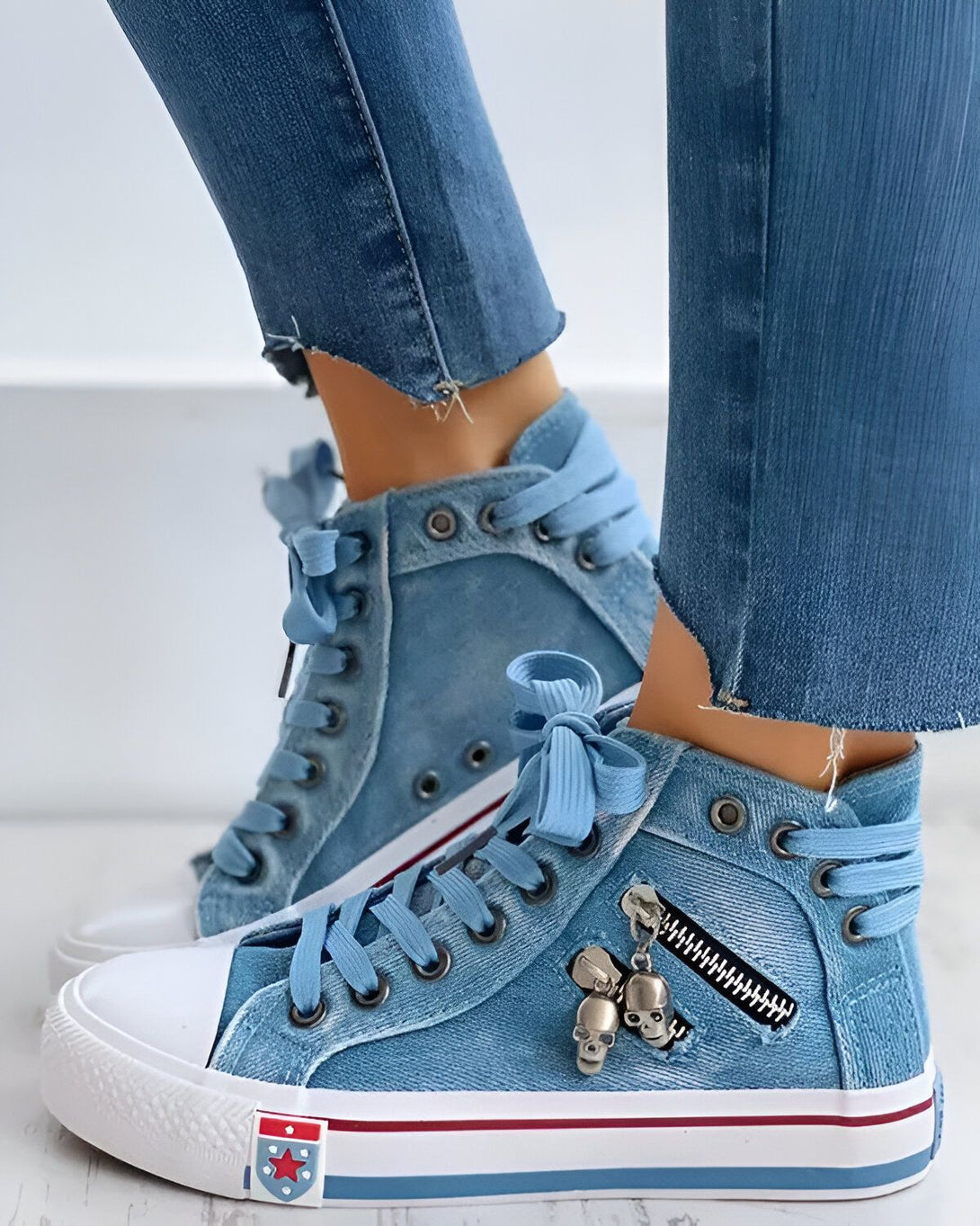 Une image d'une paire de sneakers montantes en jean bleu avec des lacets bleus et des fermetures éclair sur le côté. Il y a un pendentif en forme de tête de mort sur la fermeture éclair. La personne porte un jean bleu troué.