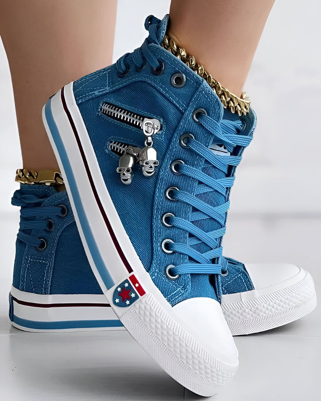 Une paire de sneakers montantes bleues avec des fermetures éclair sur le côté et des pendentifs en forme de tête de mort. Les sneakers sont portées par une femme qui porte également une chaîne dorée autour de la cheville.