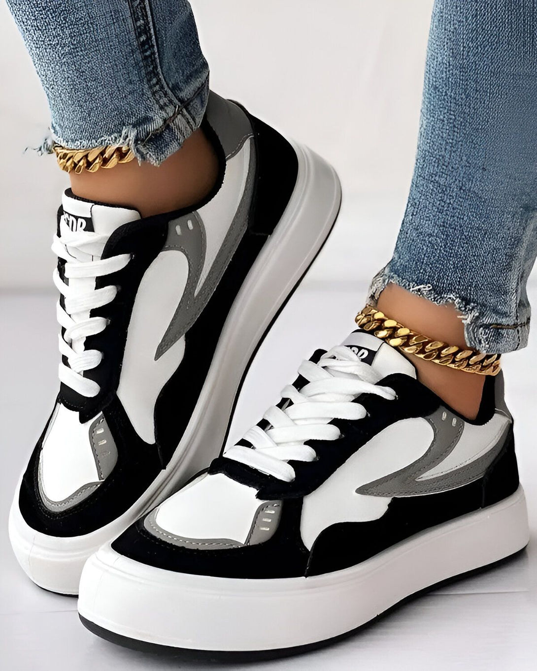 Une image d'une paire de baskets noires et blanches avec des lacets blancs. Les baskets sont portées par une femme qui porte un jean bleu et des chaussettes blanches. La femme est debout sur un sol en béton.