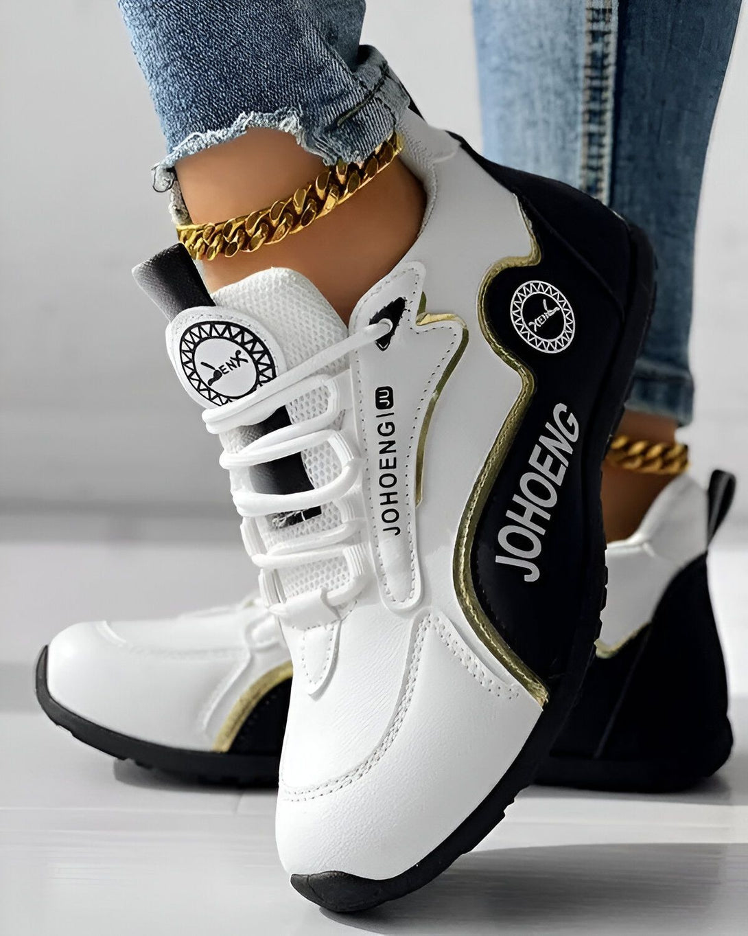 Une paire de baskets blanches et noires avec une semelle épaisse. Les baskets sont lacées et ont un logo doré sur le côté. Le pied droit porte une chaîne de cheville en or.