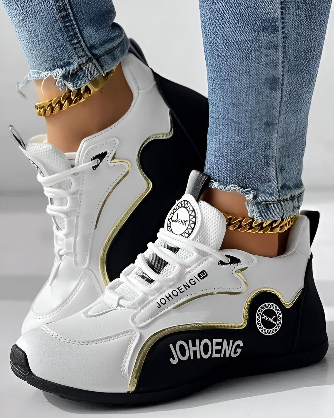 Une paire de baskets blanches et noires avec des lacets dorés. Les baskets sont portées par une femme qui porte un jean bleu et un haut blanc. La femme est debout sur un sol en béton.