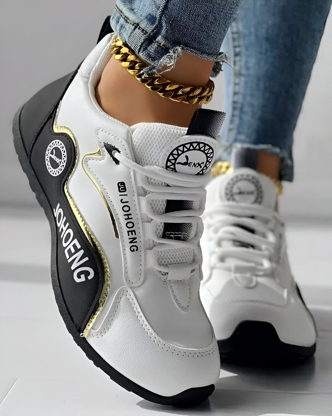 Une image d'une paire de baskets blanches et noires avec une semelle épaisse. Les baskets sont lacées et ont un logo doré sur le côté. Le pied gauche est visible avec un jean bleu et une cheville avec un bracelet doré.