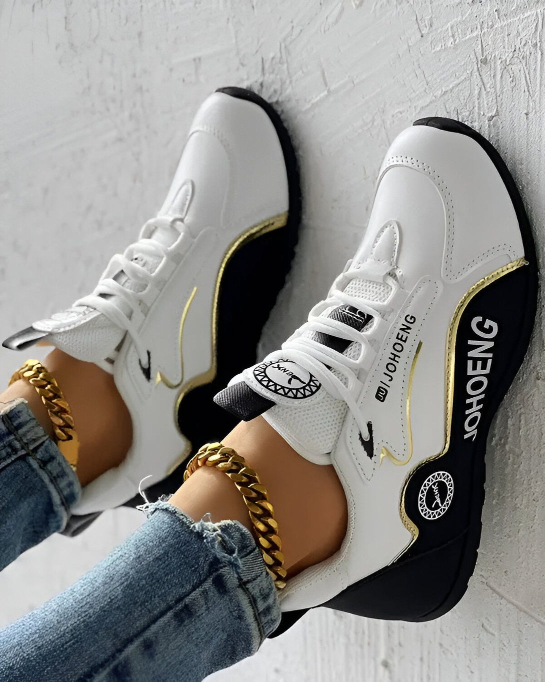 Une paire de baskets blanches et noires avec une semelle épaisse. Les baskets sont lacées et ont un logo doré sur le côté. Une personne porte les baskets avec un jean bleu et une cheville dorée.