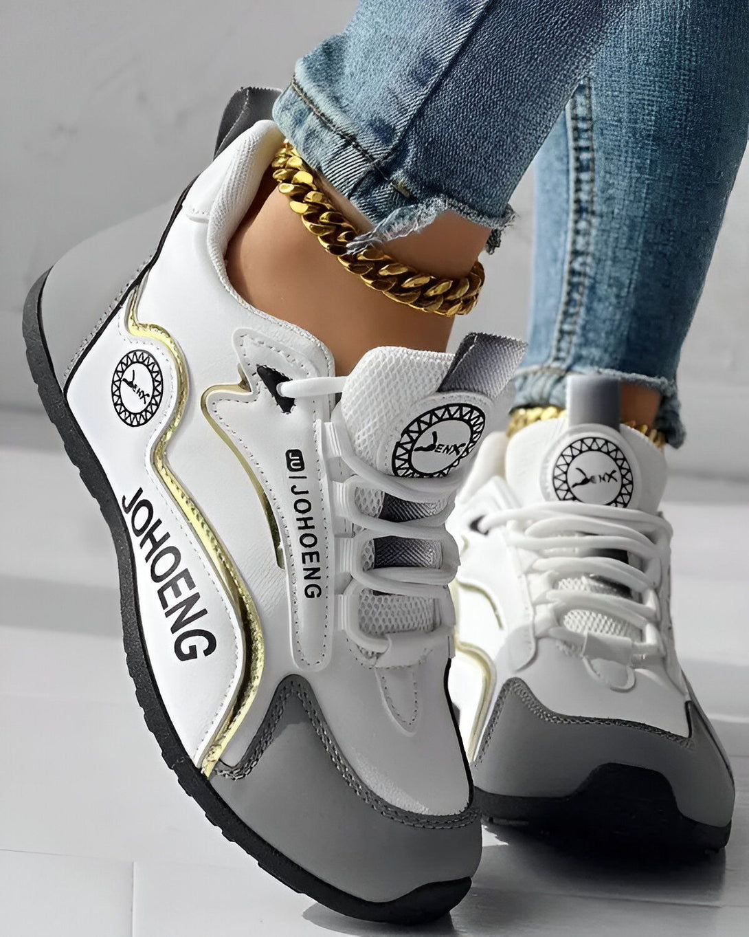 Une image d'une paire de baskets blanches et grises avec des lacets blancs. Le nom "JOHOENG" est écrit sur le côté de la chaussure. Une personne porte les baskets avec un jean bleu et une cheville dorée.