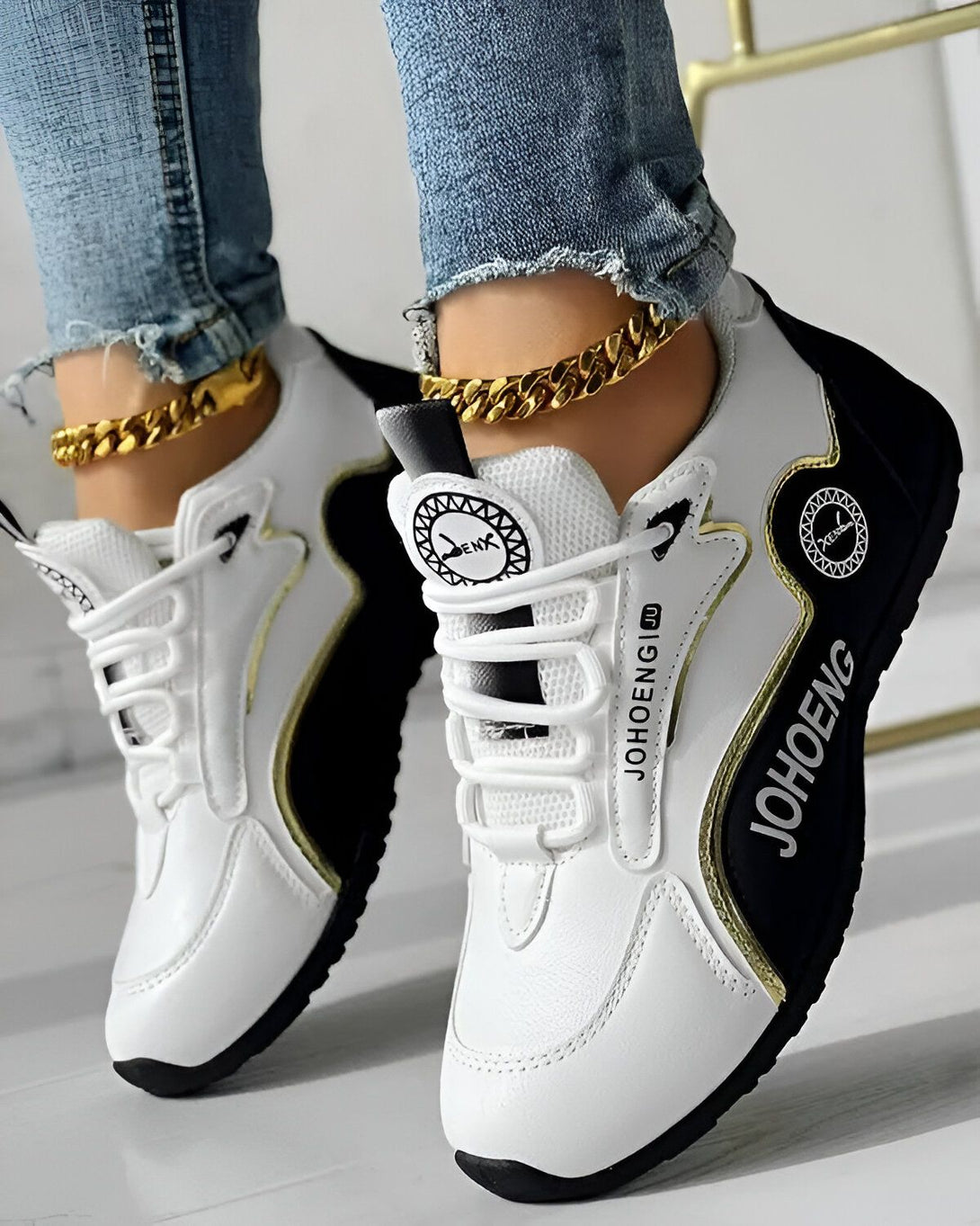 Une paire de baskets blanches et noires avec des lacets dorés. Les baskets sont portées par une femme qui porte un jean bleu et un haut blanc. La femme est assise sur une chaise et l'on peut voir ses pieds.
