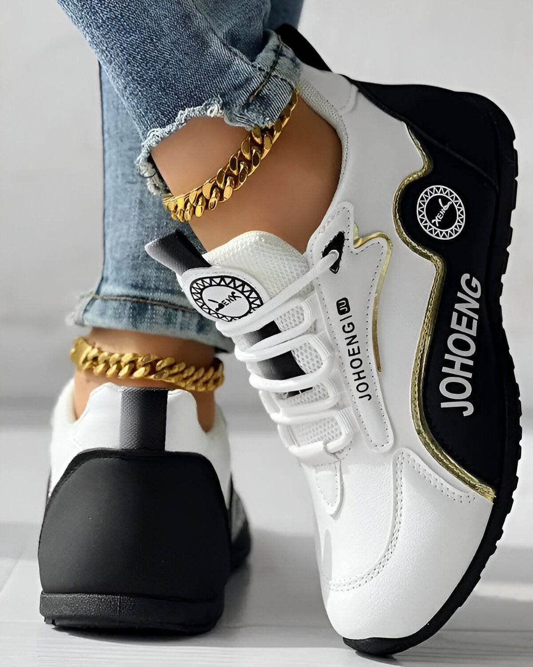 Une femme porte des baskets blanches et noires. Les baskets ont une semelle épaisse et des lacets noirs. La femme porte également un jean bleu et un bracelet doré à la cheville.