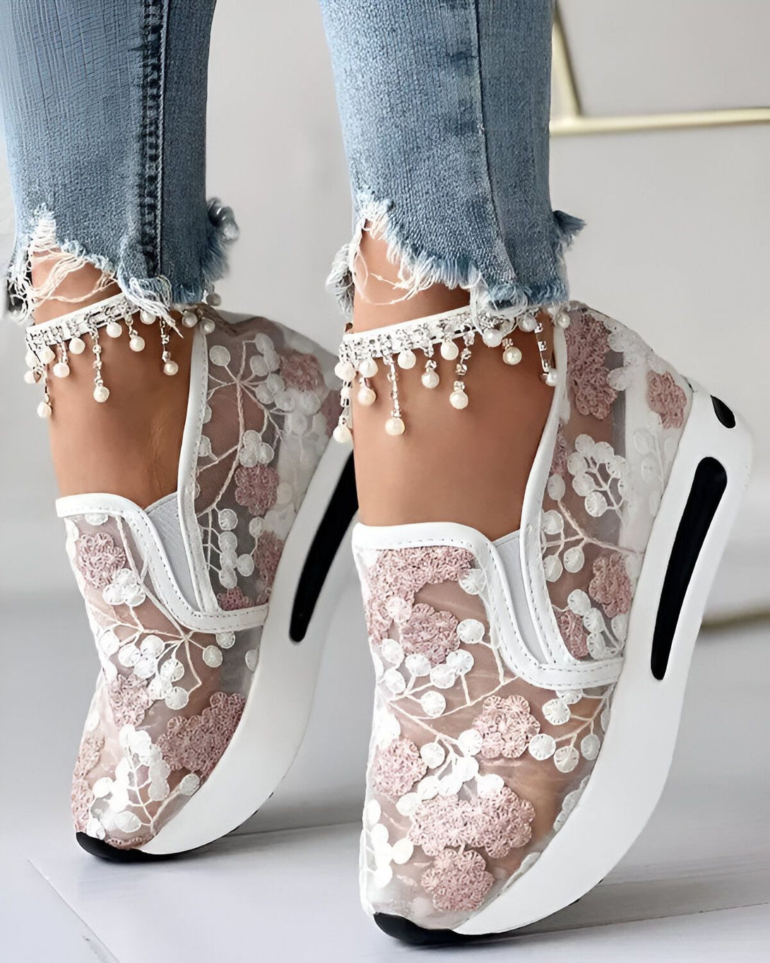 Une femme porte des baskets blanches en dentelle avec des fleurs roses. Elle porte également un jean bleu déchiré et des bijoux de cheville en perles.