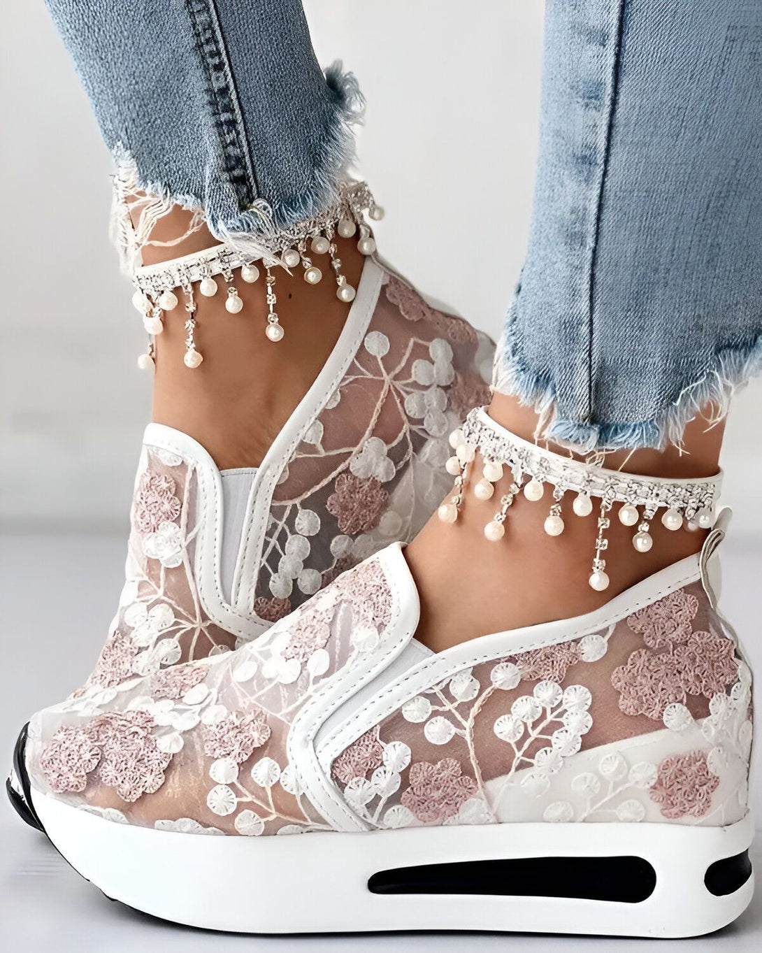 Une femme porte des baskets blanches en dentelle avec des fleurs roses. Elle porte également un jean bleu déchiré et des bracelets de cheville en perles.