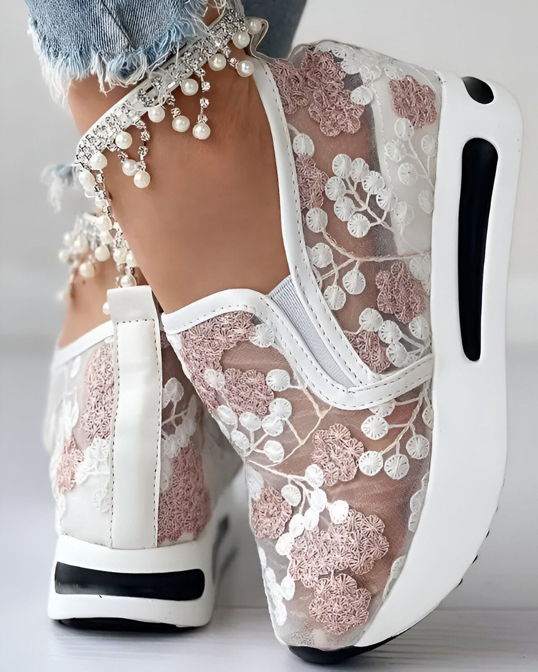 Une femme porte des baskets blanches en dentelle avec des fleurs roses. Les baskets ont une semelle épaisse et sont très confortables. La femme porte également un jean bleu et un haut blanc.