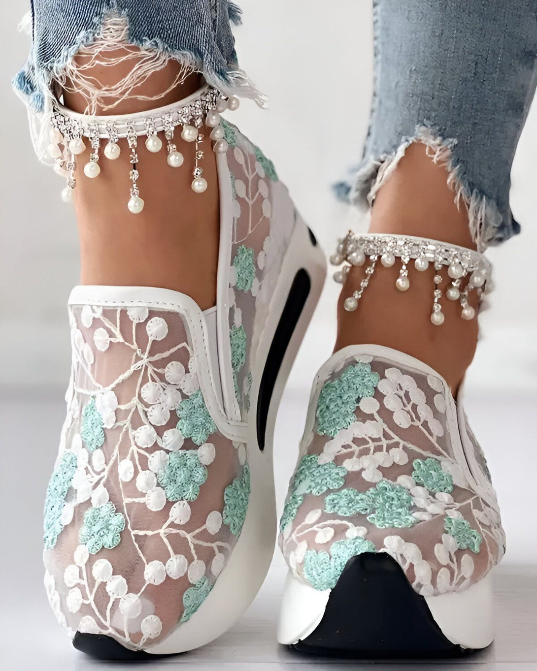 Une femme porte des baskets blanches en dentelle avec des fleurs vertes. Elle porte également des jeans bleus déchirés et des bracelets de cheville en perles.