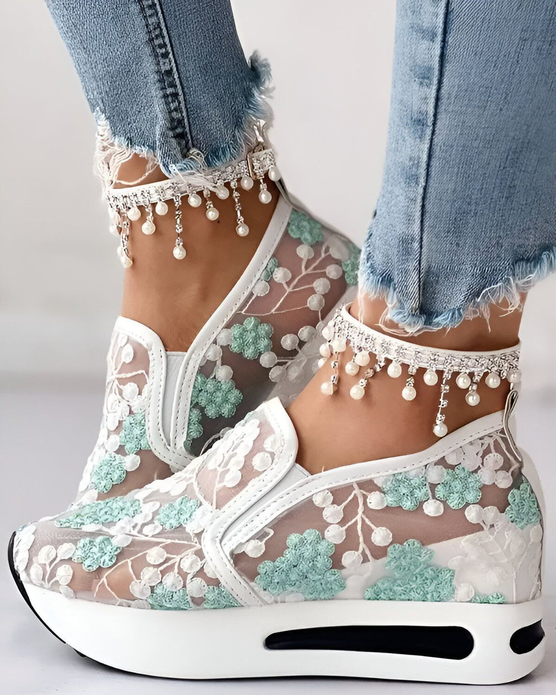 Une femme porte des baskets blanches en dentelle avec des fleurs vertes. Elle porte également des jeans bleus et des bracelets de cheville en perles.