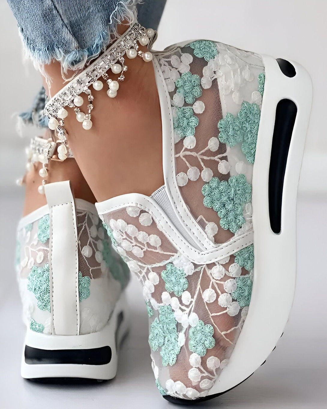 Une femme porte des baskets blanches en dentelle avec des fleurs vertes. Les baskets sont légères et semblent confortables. La femme porte également un jean bleu et un bracelet de perles à la cheville.