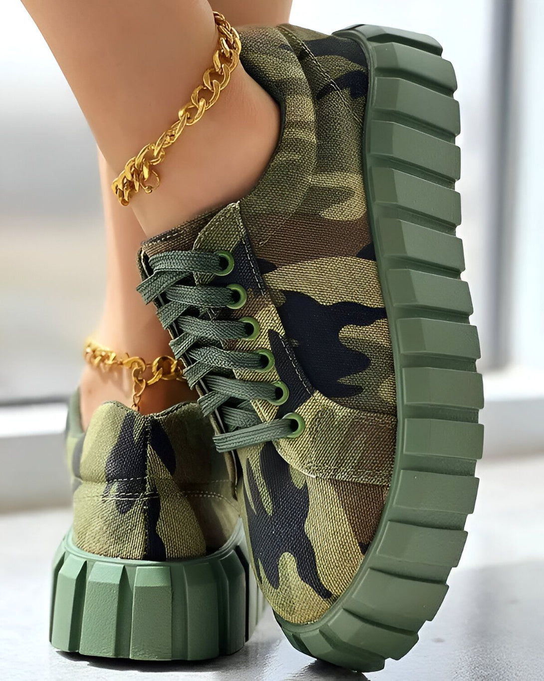 Une femme porte des chaussures de sport vert camouflage avec des lacets verts et une semelle épaisse. Elle porte également une chaîne de cheville en or.
