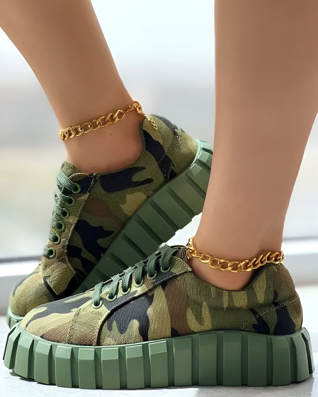 Une femme porte des baskets vert kaki à lacets avec un motif camouflage. Elle porte également des chaînes dorées aux chevilles.