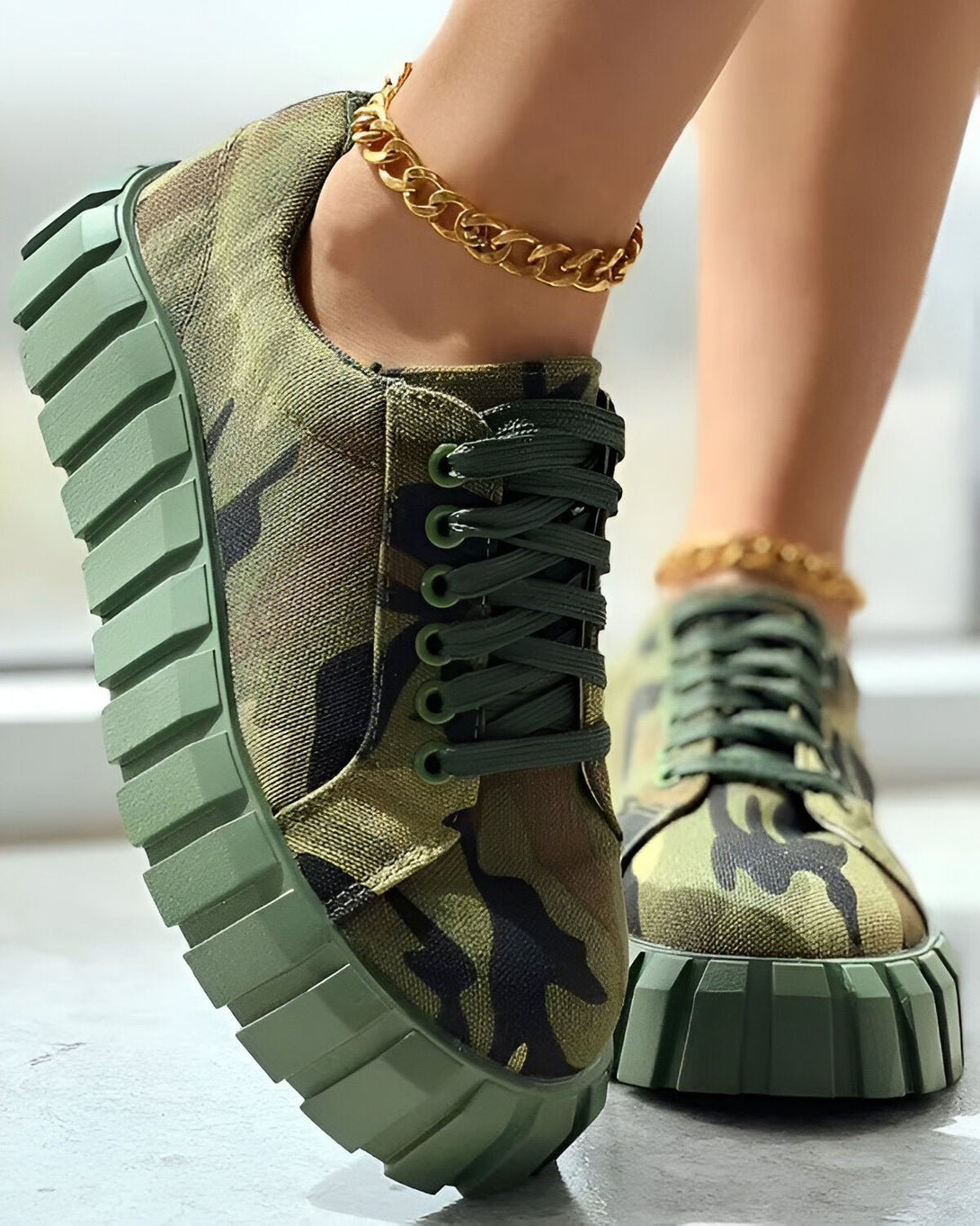 Une femme porte des baskets vertes à lacets avec un motif camouflage. Les baskets ont une semelle épaisse et crantée. La femme porte également une chaîne dorée à la cheville.
