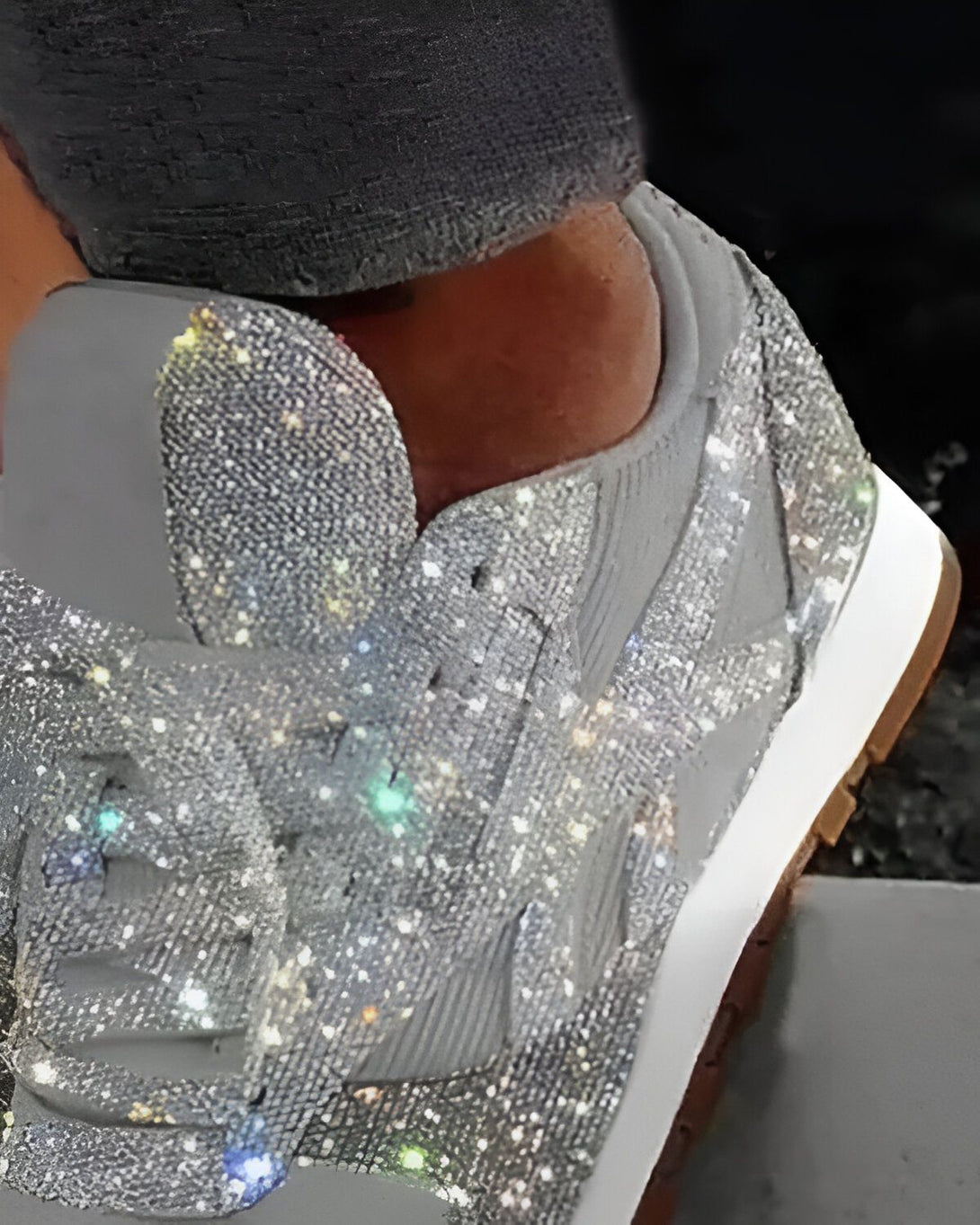 Une image d'une personne portant des baskets grises scintillantes. Les baskets sont recouvertes de paillettes argentées et ont une semelle blanche. La personne porte des chaussettes noires et un pantalon gris.