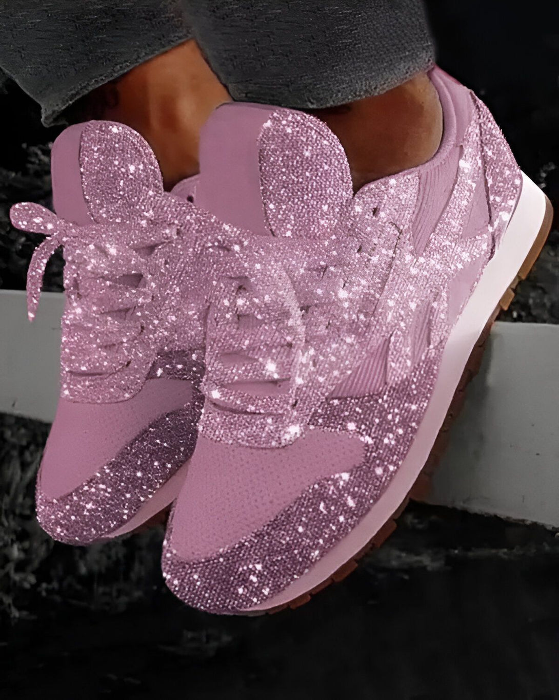 Une paire de baskets roses pailletées. Les baskets sont basses et ont des lacets. La personne qui les porte porte un jean bleu et est assise sur un muret.