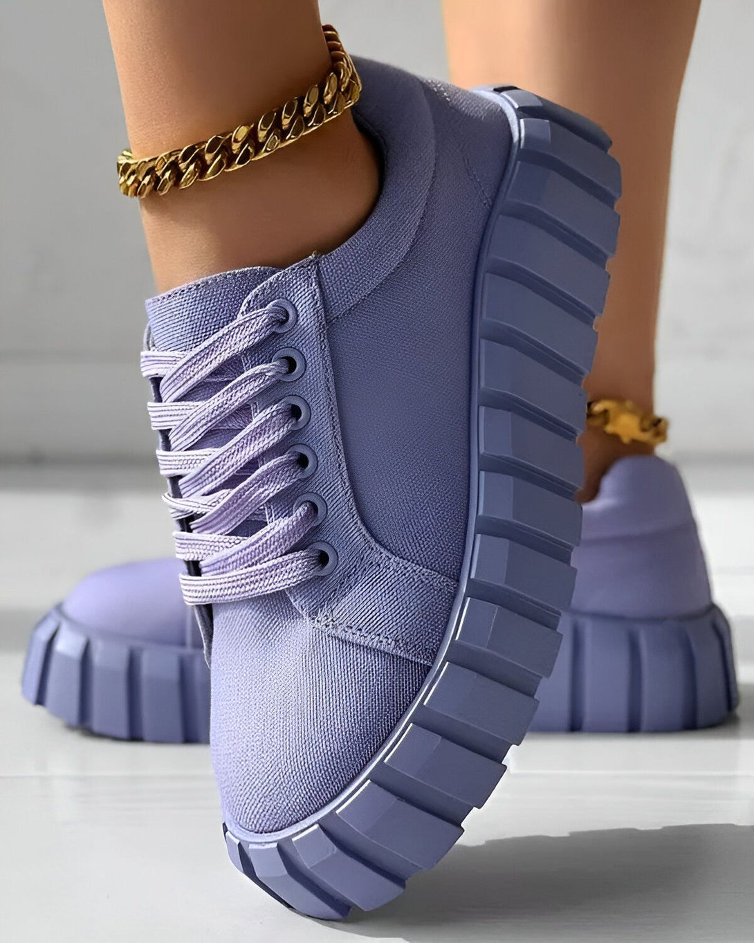 Une image d'une paire de baskets violettes avec des lacets et une semelle épaisse. Les baskets sont portées par une femme qui porte également une chaîne en or autour de la cheville. Le sol est en bois clair.