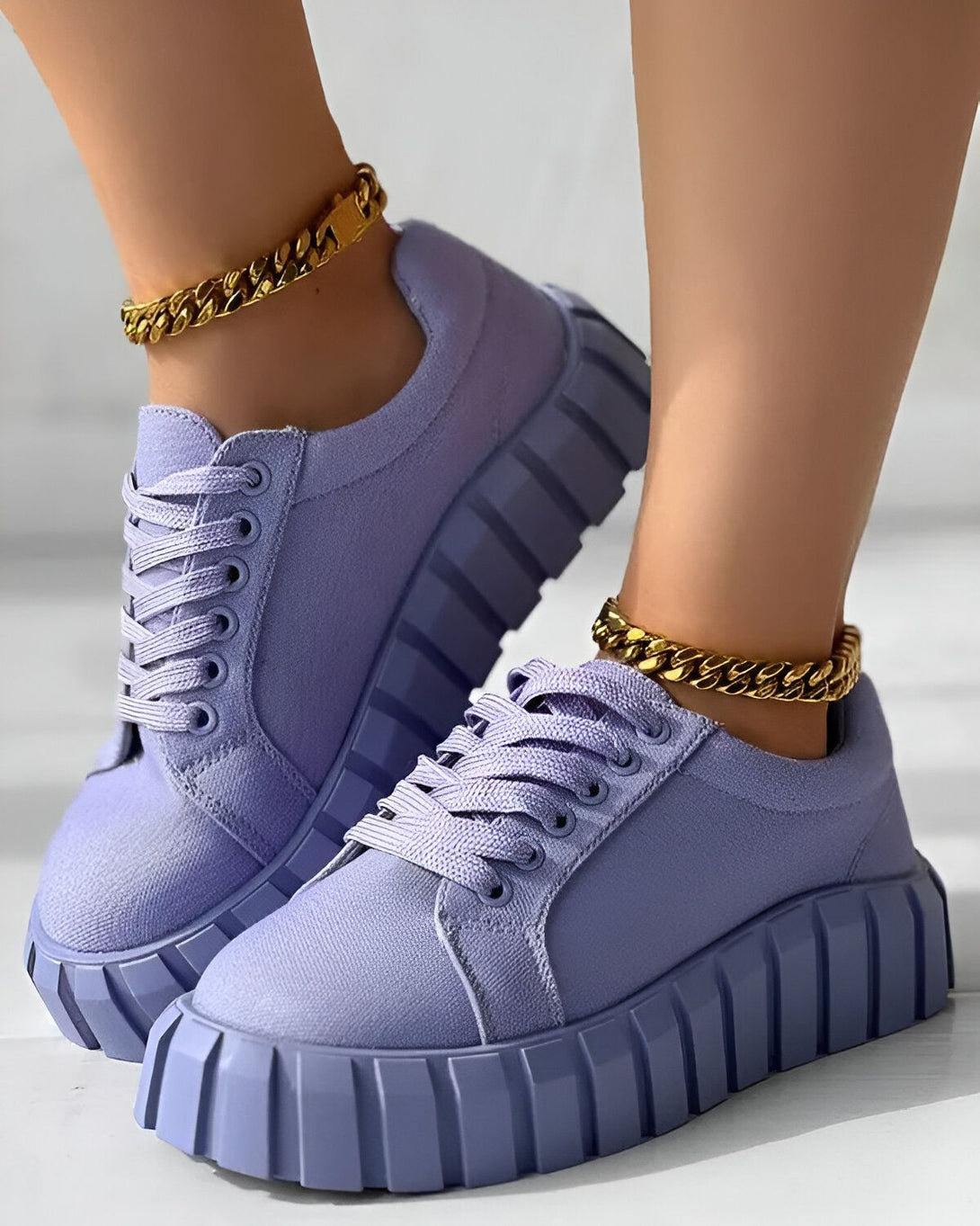 Une paire de baskets violettes avec des lacets blancs et une semelle épaisse. Les baskets sont portées par une femme qui porte également un bracelet de cheville en or.