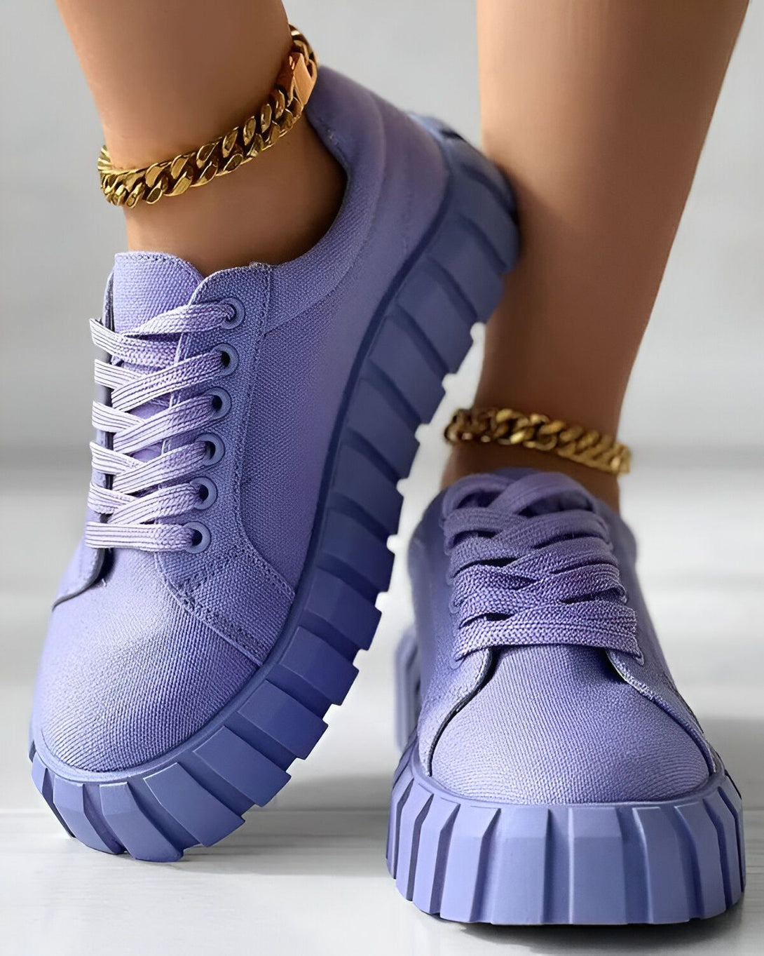 Une paire de baskets violettes avec des lacets et une semelle épaisse. Les baskets sont portées par une femme qui porte également une chaîne dorée autour de la cheville.