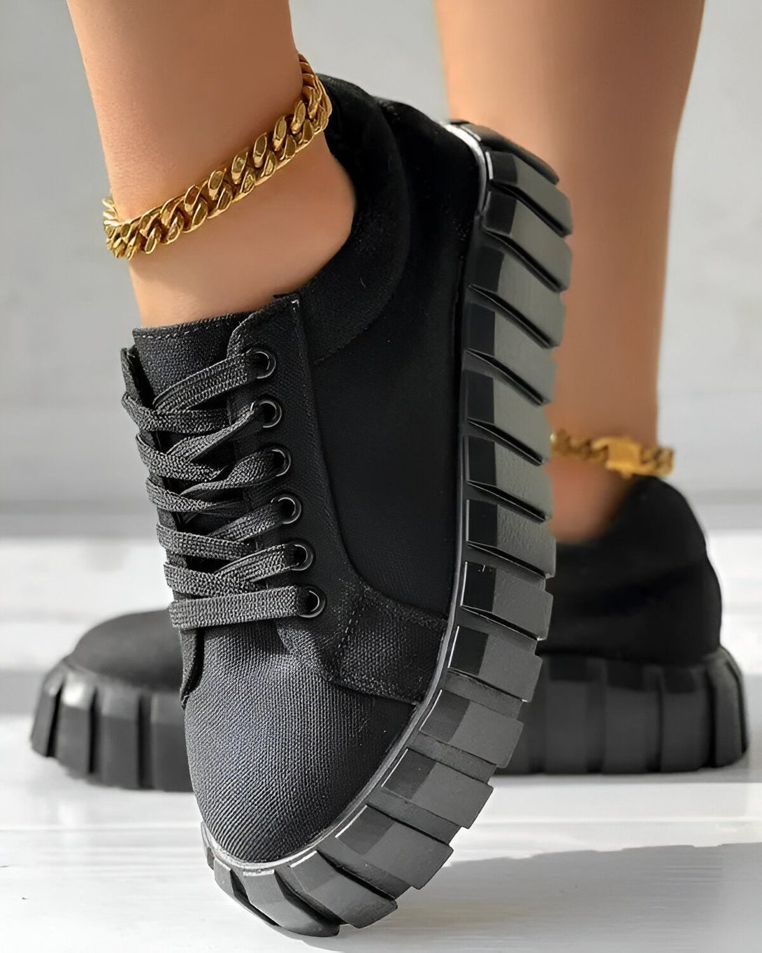 Une femme porte des baskets noires à lacets avec une semelle épaisse. Les baskets sont en toile et ont un bout rond. La femme porte également un bracelet de cheville en or.