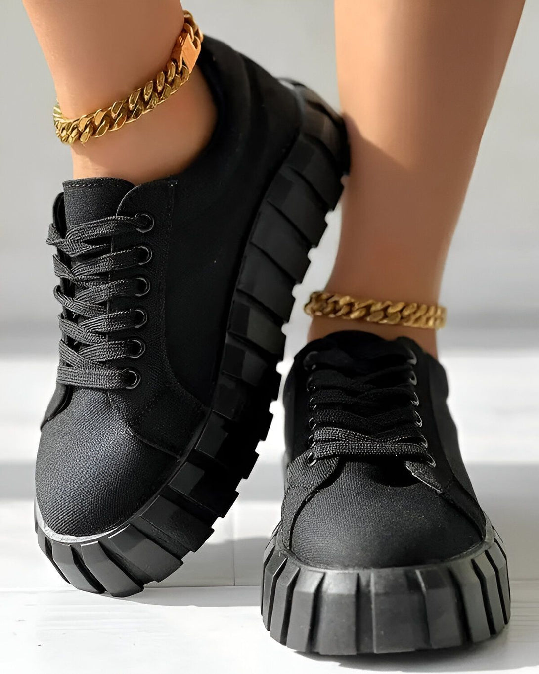 Une paire de chaussures noires à lacets avec une semelle épaisse. Les chaussures sont portées par une femme qui porte également des chaussettes noires et un pantalon noir. Le sol est en béton gris.