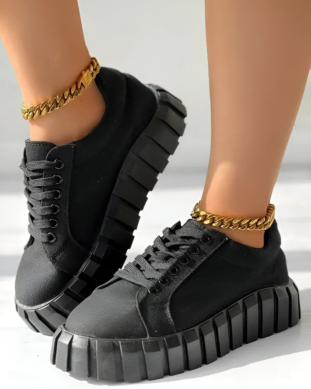 Une femme porte des baskets noires avec des lacets noirs et une semelle épaisse. Elle porte également une chaîne en or autour de la cheville.