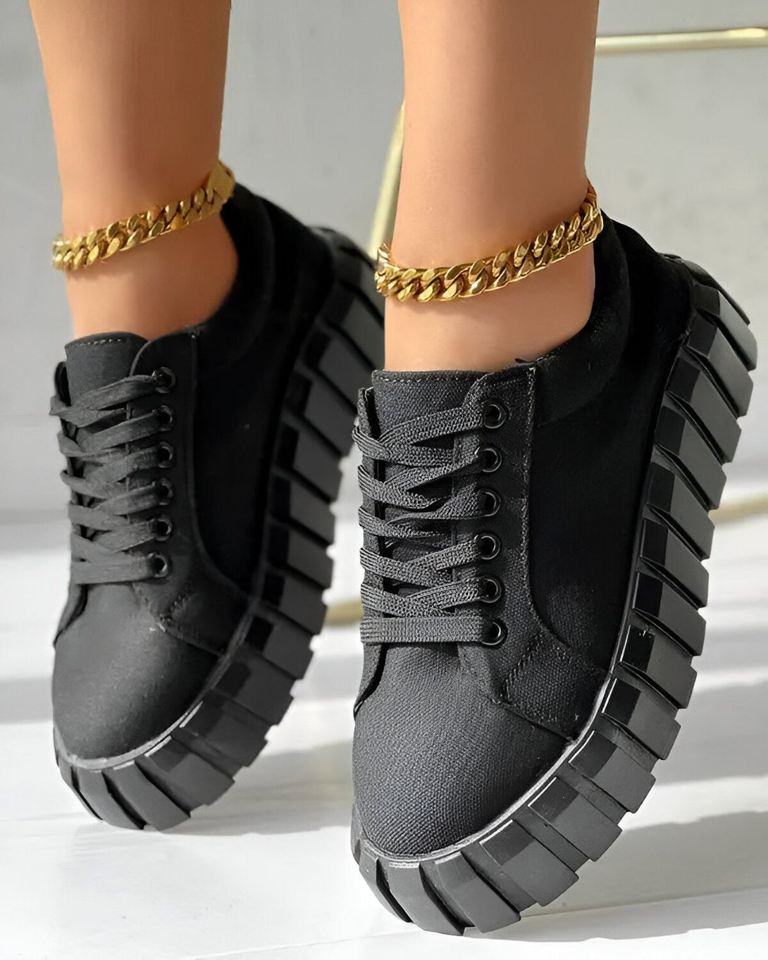 Une femme porte des baskets noires à lacets avec une semelle épaisse. Elle porte également un bracelet de cheville en or.
