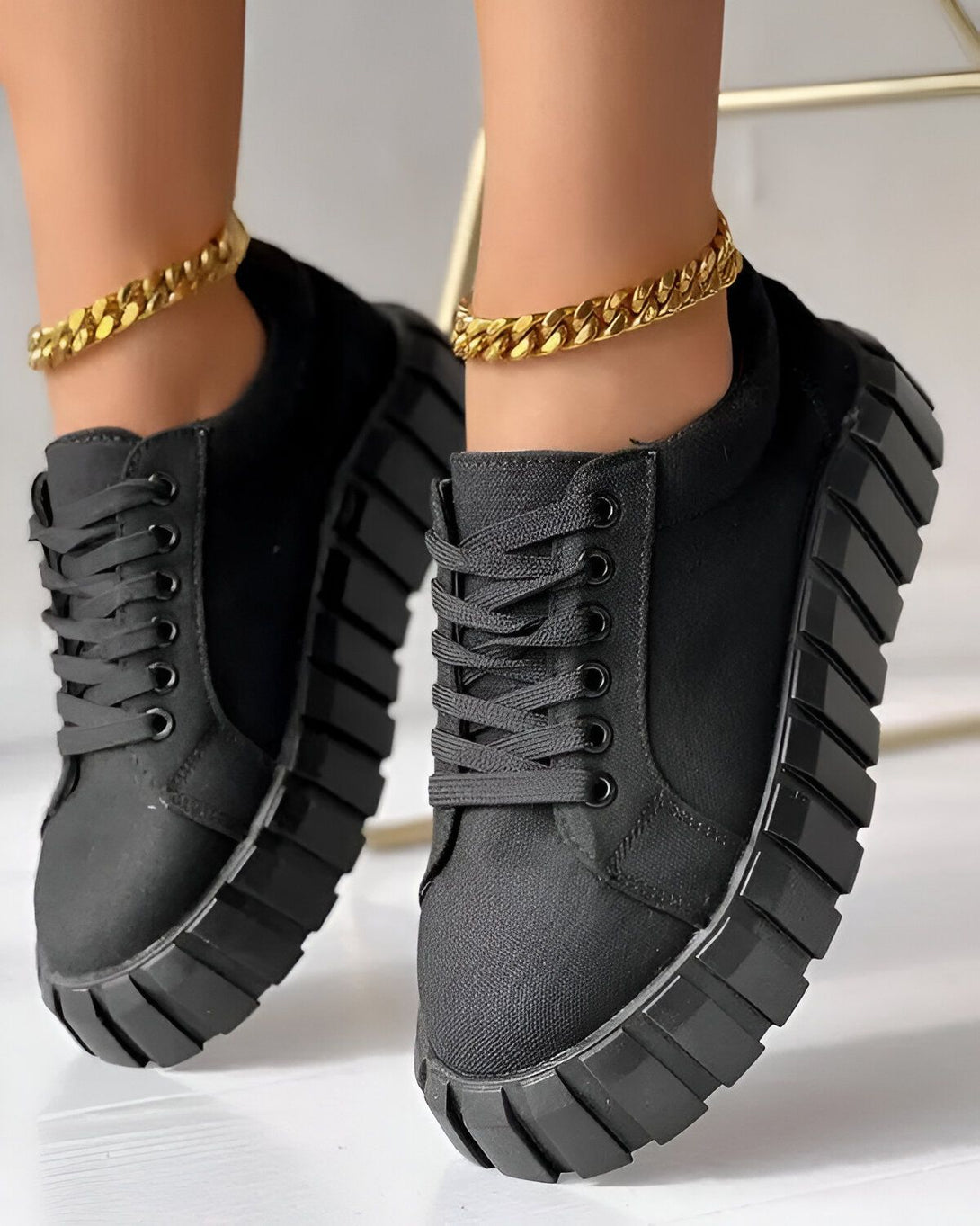 Une femme porte des baskets noires avec des lacets noirs et une semelle épaisse. Les baskets sont faites d'un matériau en toile et ont un design chunky. La femme porte également un pantalon noir et un haut blanc. Elle est assise sur une chaise et l'on peut voir ses pieds.