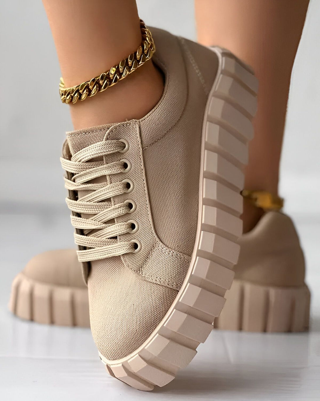 Une image d'une paire de chaussures beiges avec des lacets beiges et une semelle beige. Il y a une chaîne en or autour de la cheville du pied gauche.